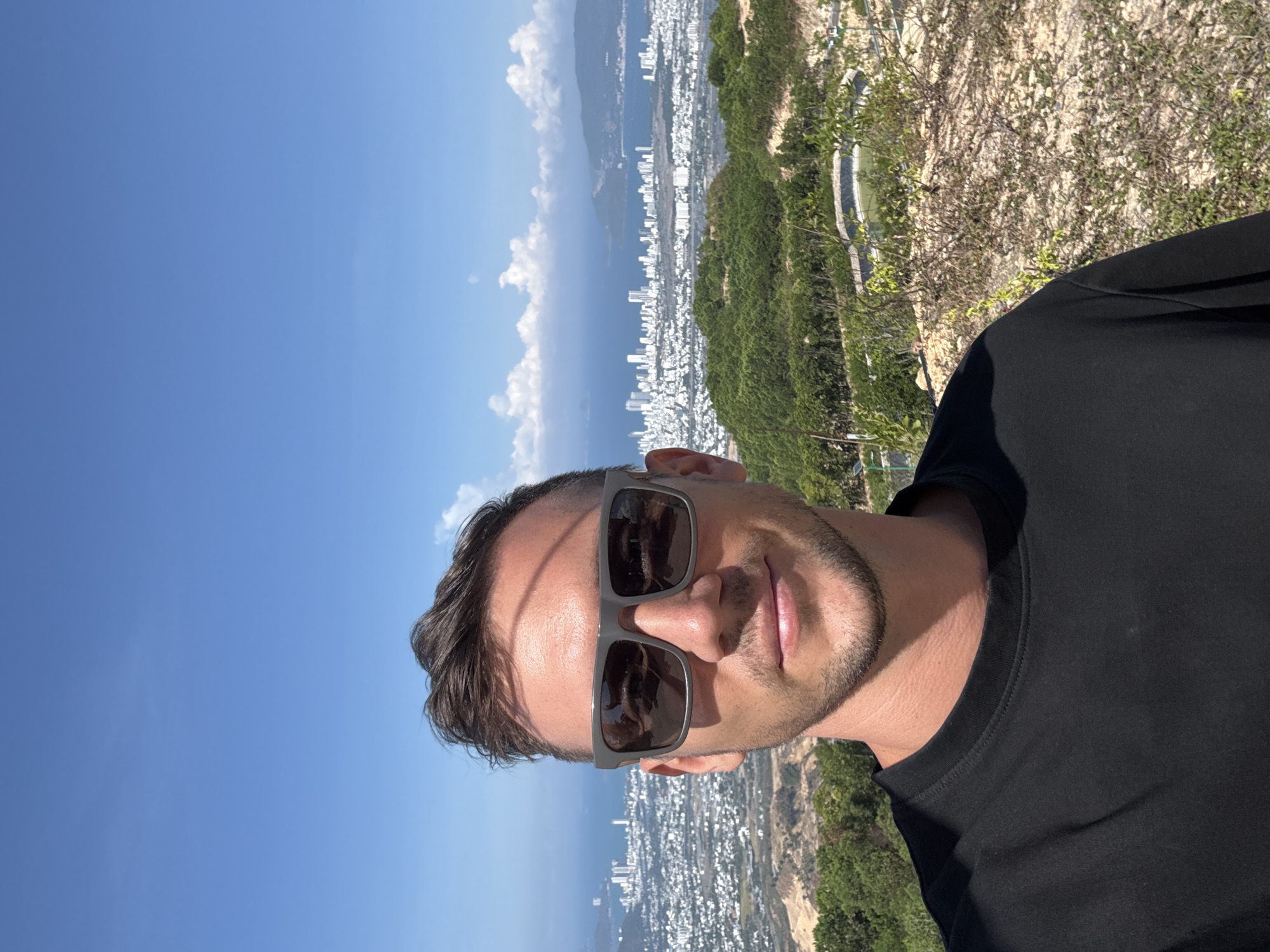 A medium shot of a man wearing sunglasses and a black t-shirt is oriented vertically. The backdrop consists of a city with numerous white buildings situated along a coastline with a body of water, possibly the ocean or a bay. There are patches of green vegetation, likely hills or mountains, surrounding the city. The sky is blue with scattered white clouds. The location may be Diên Khánh, Vietnam.