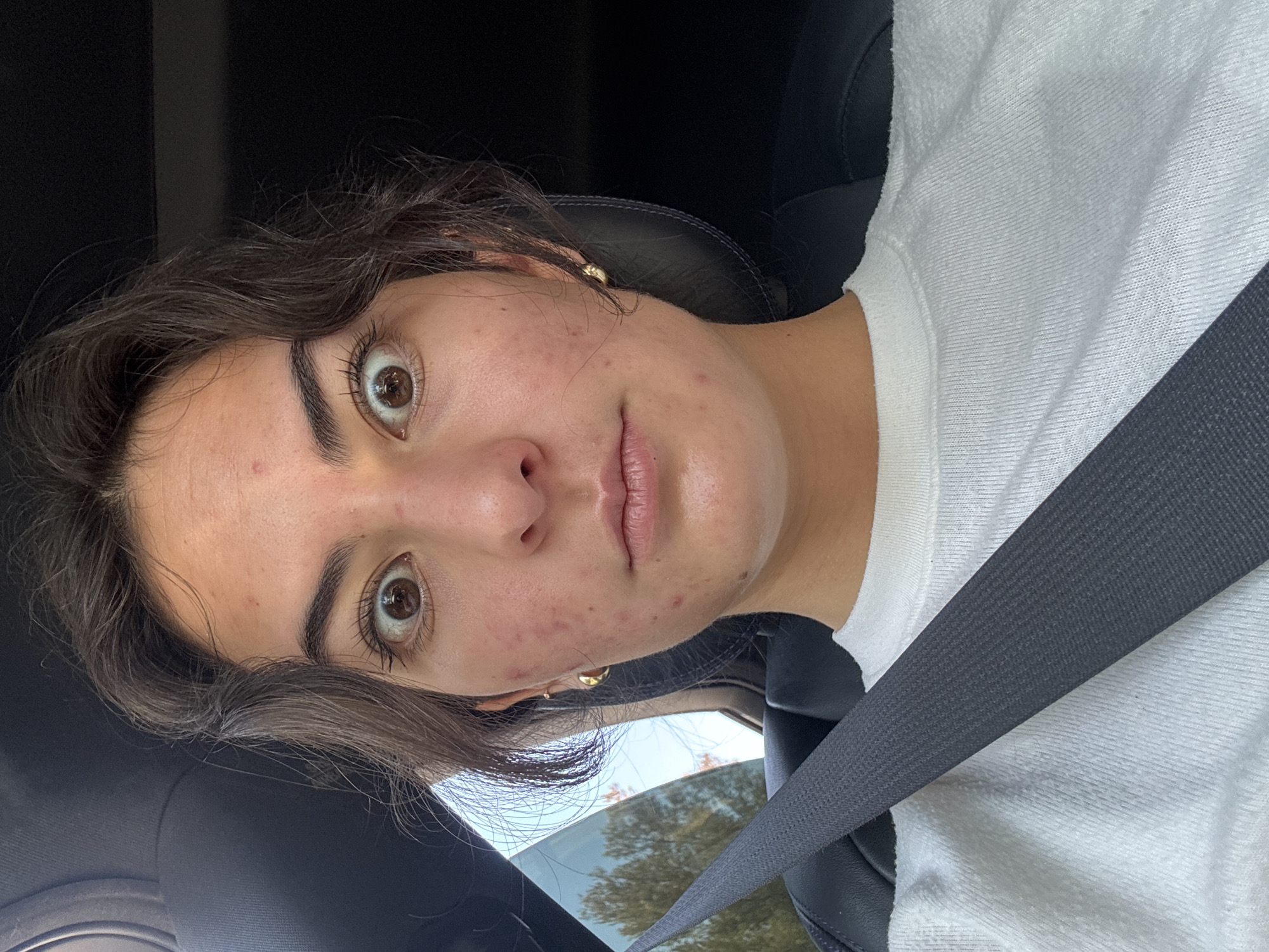 A woman with brown hair and eyes, wearing a white shirt and a seatbelt, is in a car. The background visible through the car window suggests an outdoor environment with trees. The scene does not provide enough information to confirm the location in the city of Puebla, Mexico.