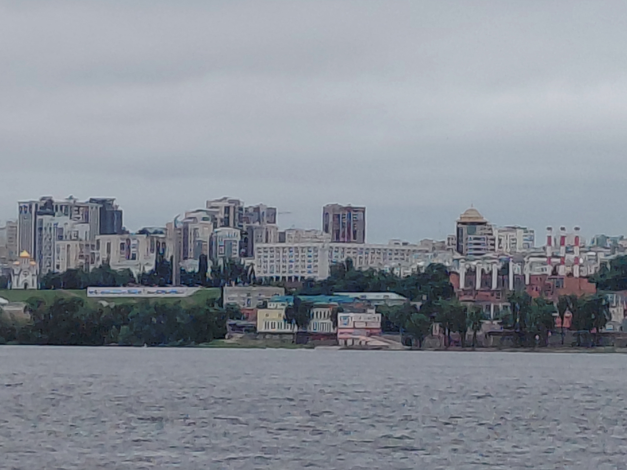 A wide shot captures the city of Rozhdestveno, Russia, from across a body of water under a cloudy sky. The city skyline is dominated by a mix of modern high-rise buildings and older, lower structures, interspersed with trees. In the foreground, the water's surface is textured with ripples. The buildings on the waterfront range from multi-story apartment complexes to smaller, more industrial-looking buildings, with various architectural styles visible. A notable structure with a dome stands out on the right side of the skyline. A strip of green trees runs along the edge of the city, separating the water from the buildings. The overall impression is one of a developed urban area situated alongside a natural waterway.