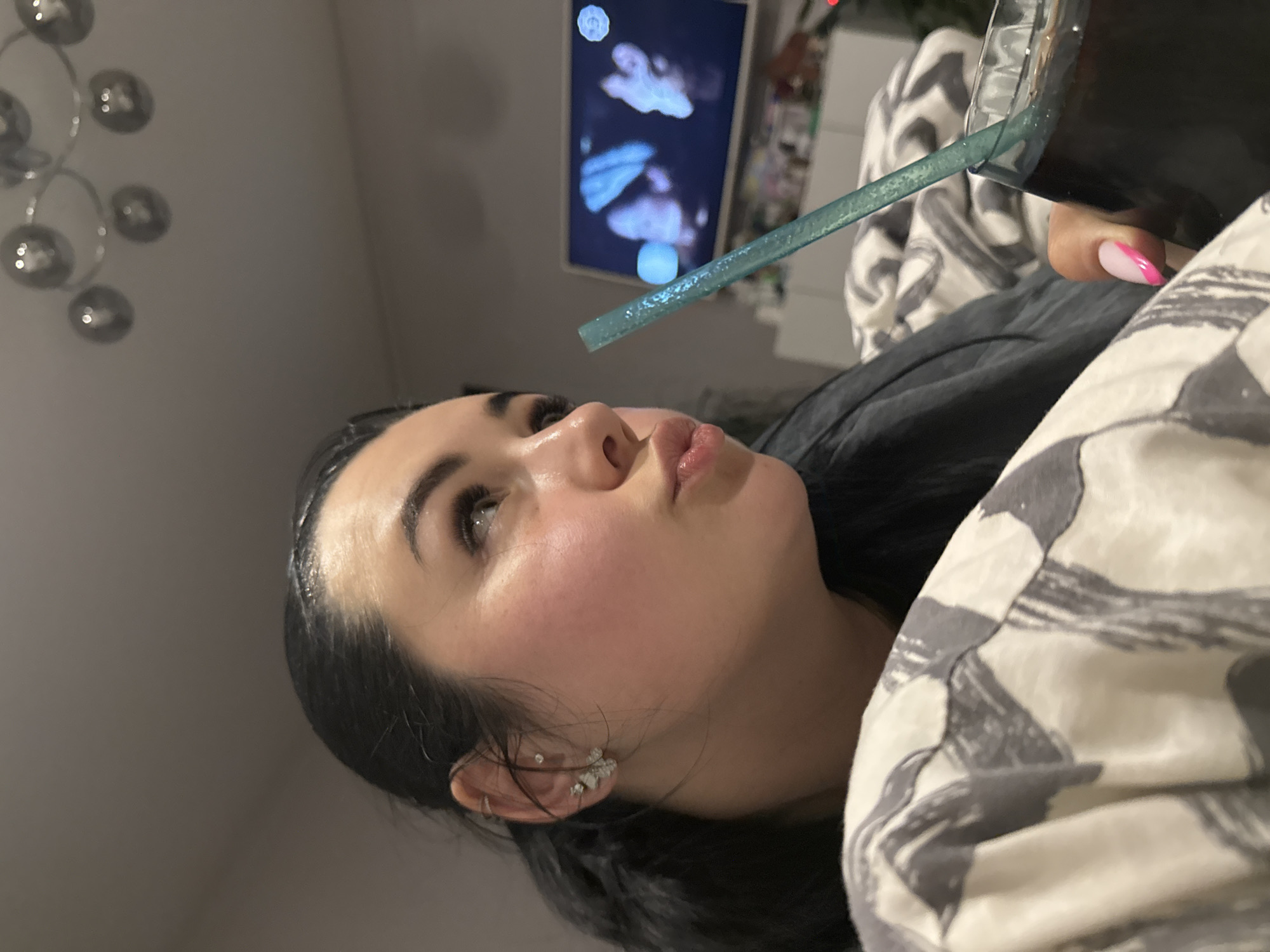 A woman is lying down, possibly in bed, with a drink in a glass and a blue straw. She has dark hair pulled back, fair skin, and is wearing earrings. Her fingernails are painted with a pink and white design. A patterned duvet covers her. In the background, a television screen displays an image of jellyfish. A chandelier is visible on the ceiling.
I cannot infer the location is in the city of Chaikovsky, Russia from the image.
