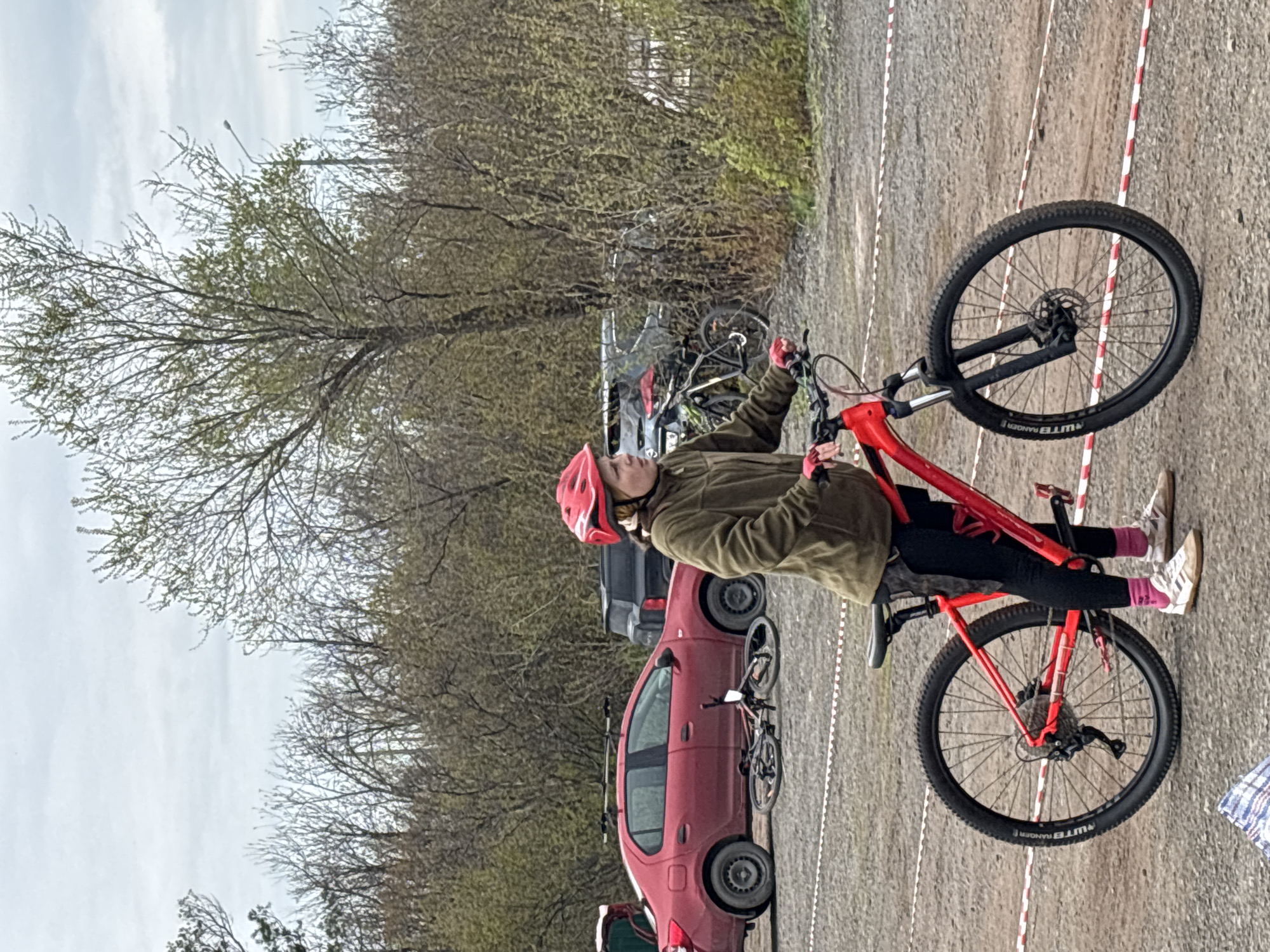 A person wearing a red helmet and an olive-green jacket is sitting on a red mountain bike. The bike is positioned on a gravel surface marked with red and white striped tape. A red car and other parked vehicles are visible in the background, along with trees. It is not possible to confirm that the location is in the city of Orenburg, Russia, as there is no geographical context or identifying information in the image.