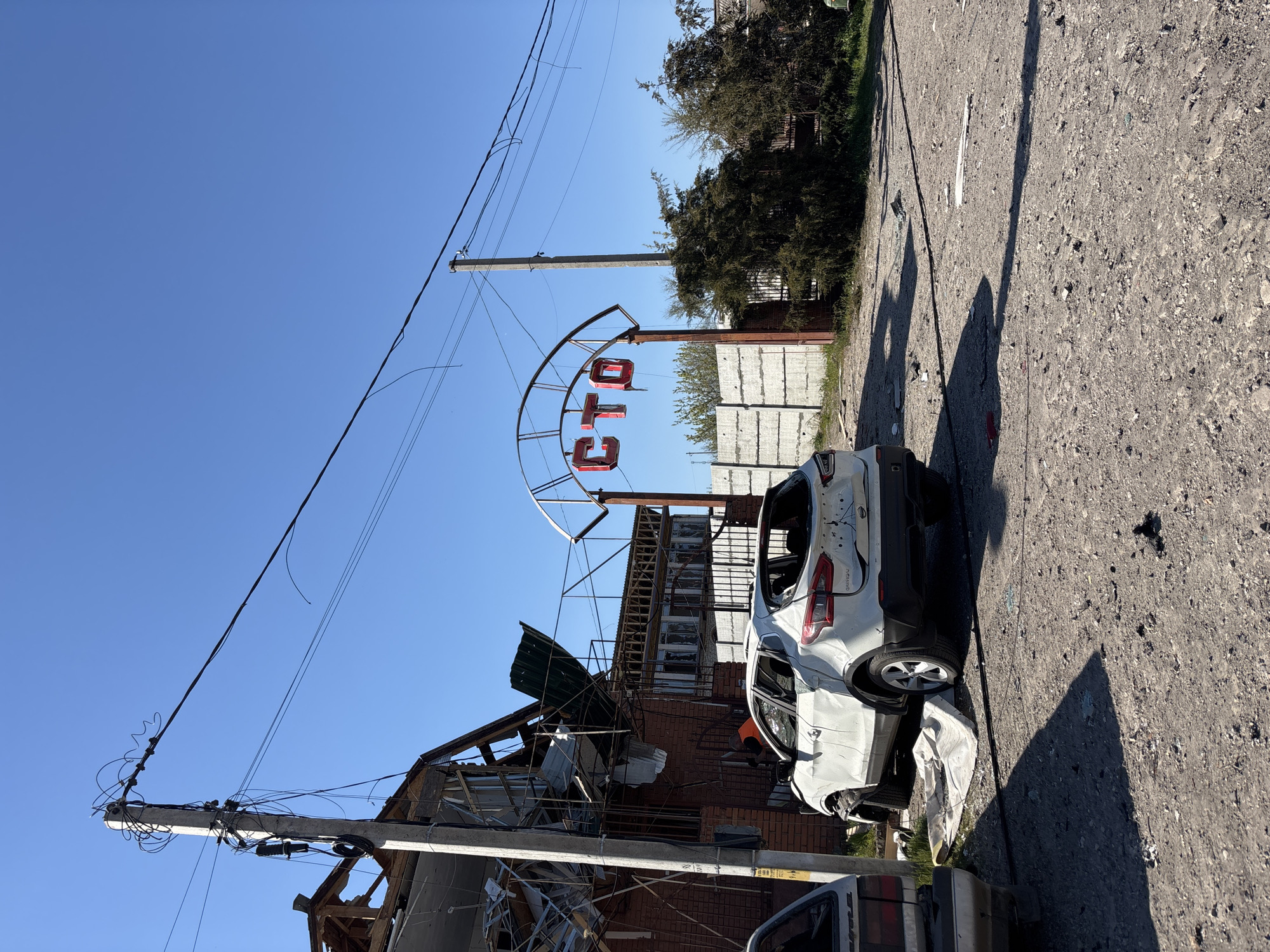The image depicts the aftermath of destruction in Pokotylivka, Ukraine. A heavily damaged white Nissan Qashqai sits on a cracked asphalt road. The vehicle has shattered windows, visible bullet holes, and significant body damage. Beside it, debris lies scattered on the ground. In the background, a brick building is severely damaged, with parts of its roof and structure collapsed. A sign with red Cyrillic letters spelling "CTO" (likely referring to a vehicle service station) stands near the building. Utility poles with dangling wires are visible. The sky is clear and blue, suggesting it is daytime. The overall scene suggests recent conflict or shelling.