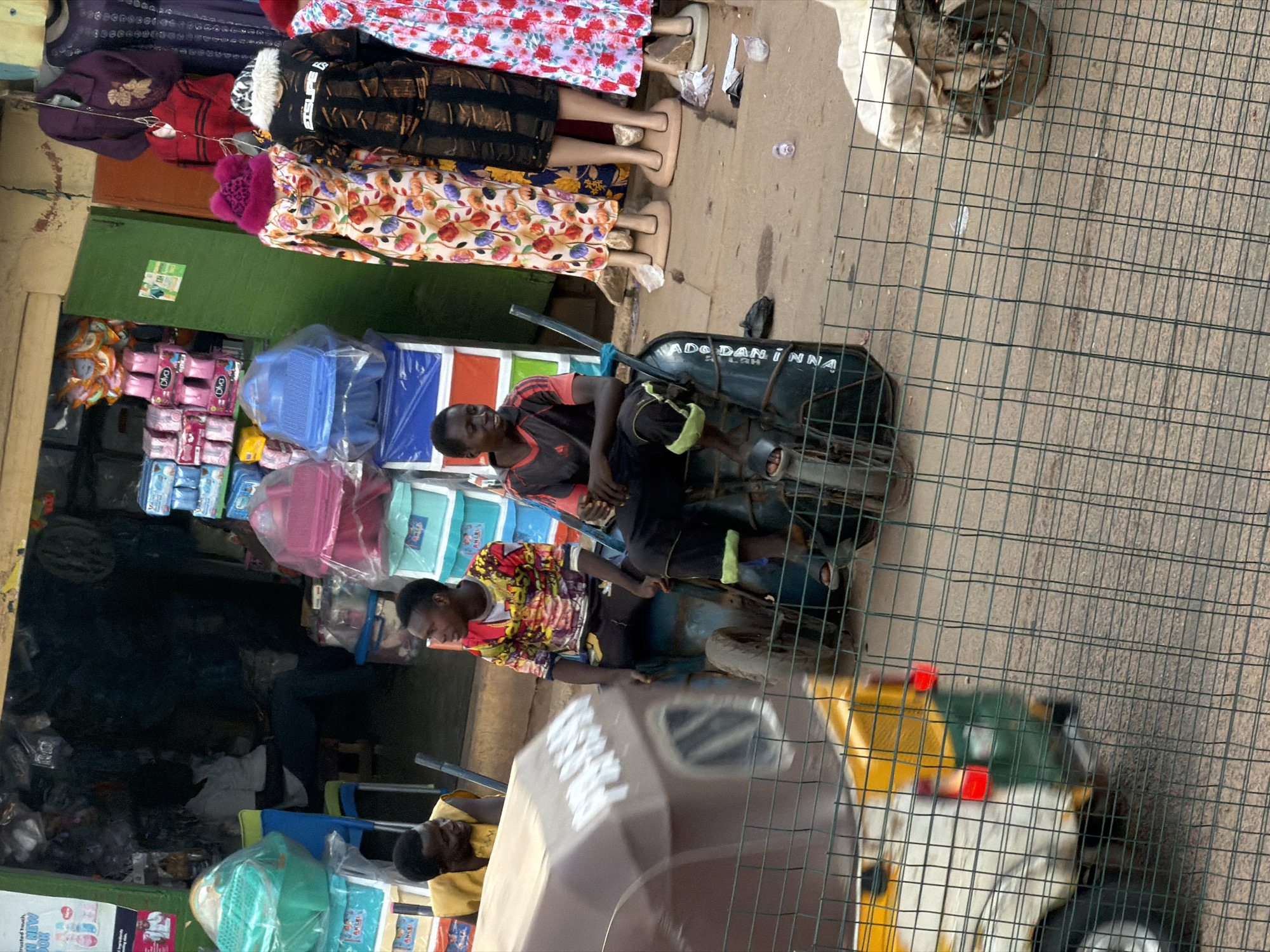 In Bukuru, Nigeria, an outdoor market scene unfolds. A brown and yellow auto-rickshaw is parked in the foreground, partially obscured by a wire fence. To the left of the rickshaw, a man in a yellow vest is visible, facing away from the camera. To the right, two young men are seated on what appears to be a cart, interacting with each other. One is wearing a colorful long-sleeved shirt, while the other sports a dark shirt with red accents. Behind them, a storefront is packed with goods, including stacks of colorful plastic containers, bags of toiletries, and clothing on mannequins. The overall impression is one of bustling commerce in a vibrant street setting.