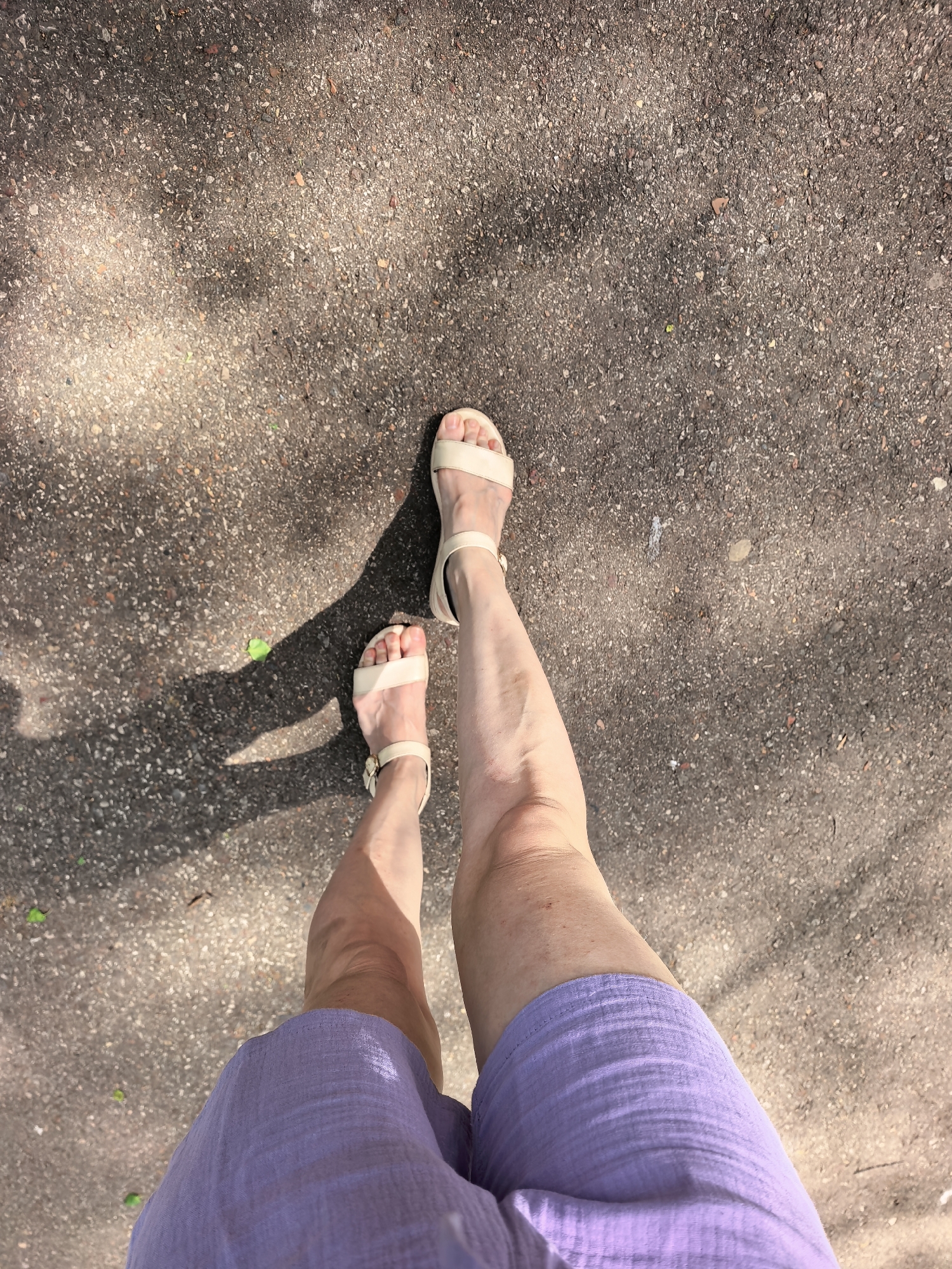 A first-person perspective shot looking down at a person's legs and feet. The person is wearing light purple shorts and beige sandals. They are standing on a paved surface that appears to be a sidewalk or street, speckled with dark spots and patches of light and shadow. The light suggests the presence of trees overhead. No specific landmarks indicate the location is Sterlitamak, Russia.