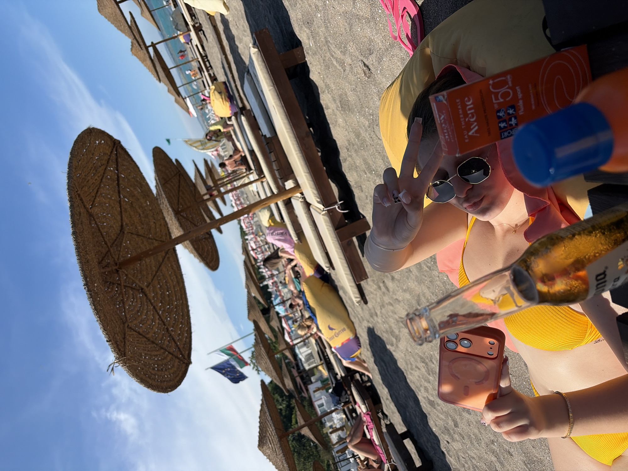 A person lies on a yellow beanbag on a sandy beach, under a thatched umbrella. The person is wearing a yellow bikini and sunglasses, and holds a smartphone in their left hand. Next to them is a bottle of beer and a box of Avene sunscreen. In the background, other people are relaxing on beach chairs, and more umbrellas are visible. Flags of Bulgaria and the European Union are planted in the sand behind the beach chairs. The scene is illuminated by bright sunlight. The location is the central beach in Burgas, Bulgaria.