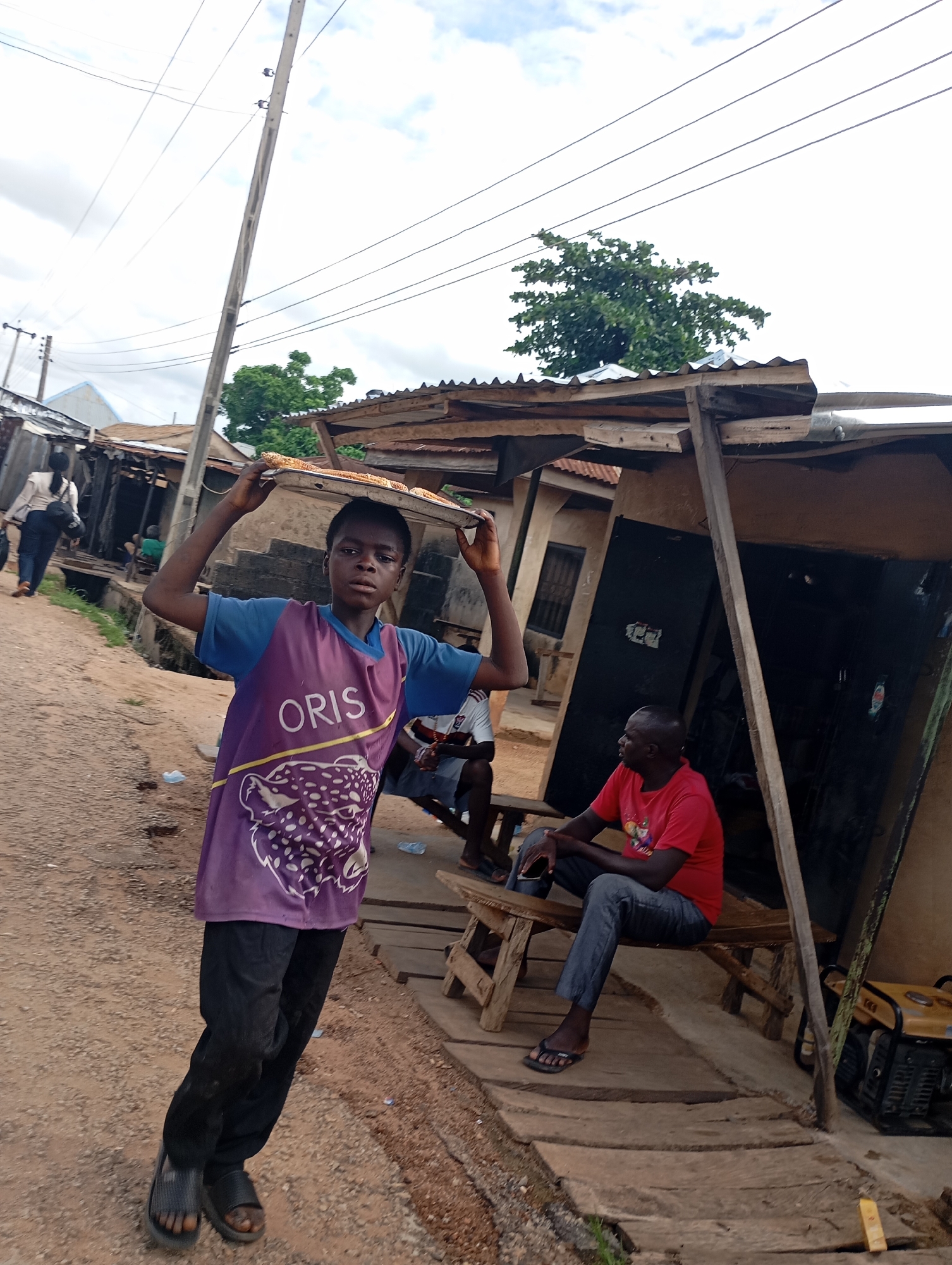 A young boy, wearing a purple t-shirt with "ORIS" printed on it and black pants, walks on a dirt road in Shendam, Nigeria, carrying a tray of food on his head. He is looking towards the camera. To his right, a man in a red t-shirt and gray pants sits on a wooden bench outside a corrugated metal structure. The structure appears to be a small shop or stall. In the background, a power line pole with wires runs overhead. Other people and structures are visible in the distance, suggesting a busy street or market area.
