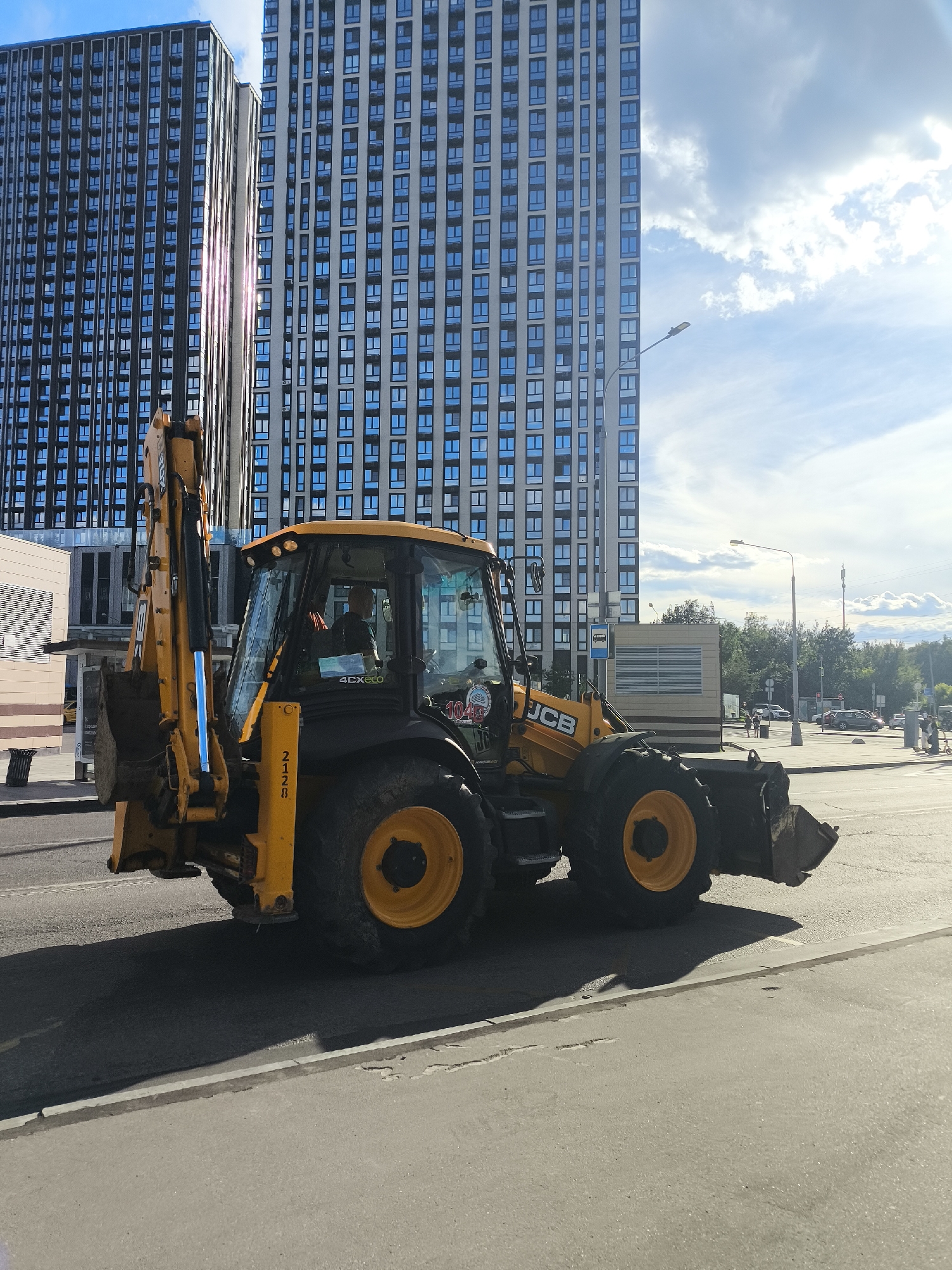 A bright yellow backhoe loader, manufactured by JCB, is parked on an asphalt road in front of two tall, modern apartment buildings with numerous blue-tinted windows. The sky above is a mix of blue and white clouds, suggesting it is daytime, possibly late afternoon given the sun's angle and the long shadows cast by the vehicle. The backhoe has a driver visible inside the cab, wearing a dark shirt. There are various decals on the vehicle, including "4CXeco" and "JCB." A numerical decal on the side of the vehicle reads "2128". A lamppost is visible to the right of the backhoe, and further in the background, there are trees, other vehicles, and a bus stop shelter. The overall scene appears to be a street in an urban environment, possibly during a construction or road maintenance period, given the presence of heavy machinery.