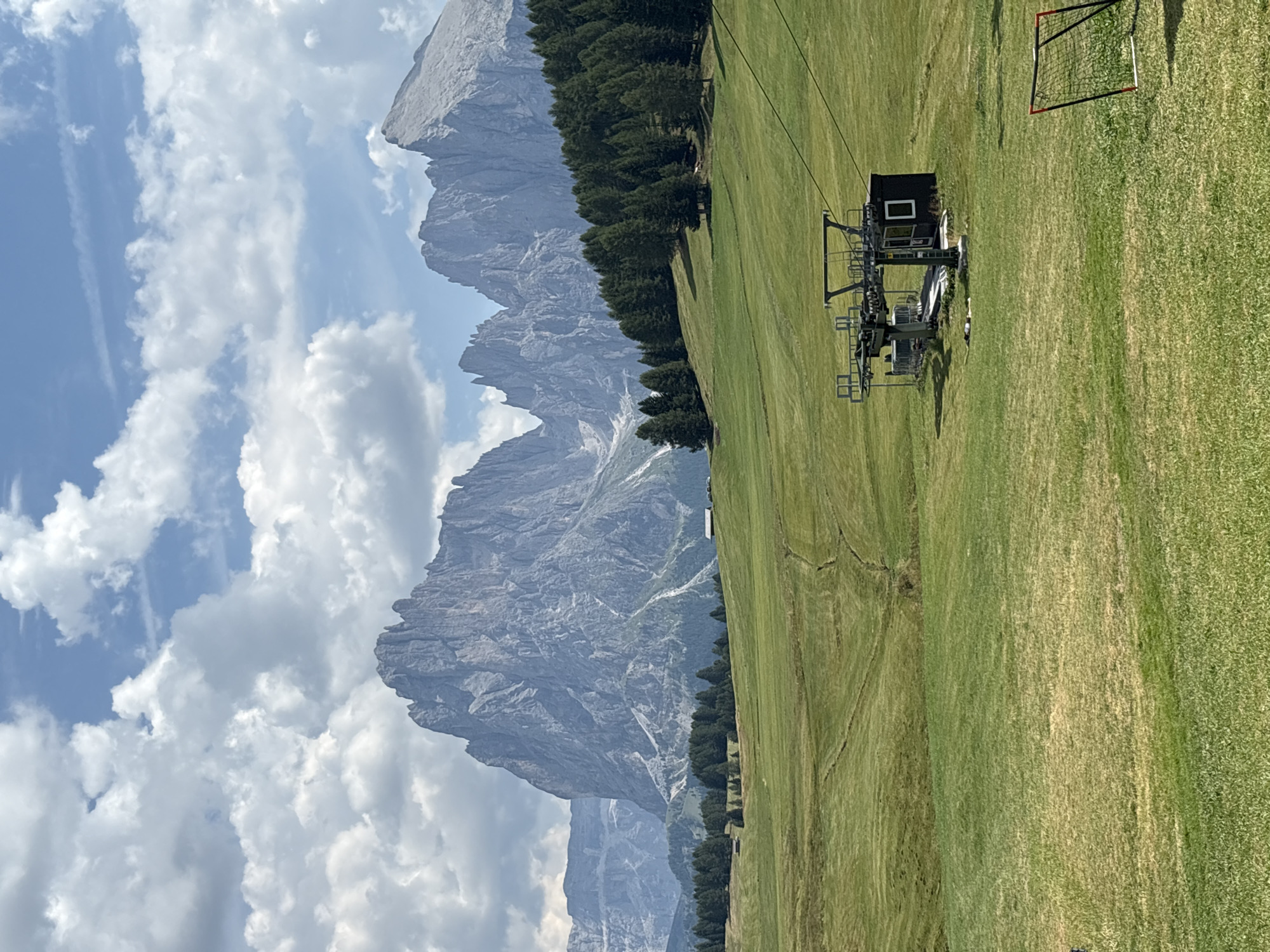 The image depicts a scenic view of the Dolomite mountains in Kastelruth - Castelrotto, Italy, on a cloudy day. In the foreground, a lush green meadow slopes gently, dotted with scattered trees and a small ski lift station with a dark building. Further back, a dense forest of evergreen trees lines the base of the imposing mountain range. The mountains themselves are rugged and rocky, with sharp peaks and dramatic geological formations. The sky is a mix of blue patches and white, fluffy clouds. There is a small soccer goal visible in the lower right corner of the image.
