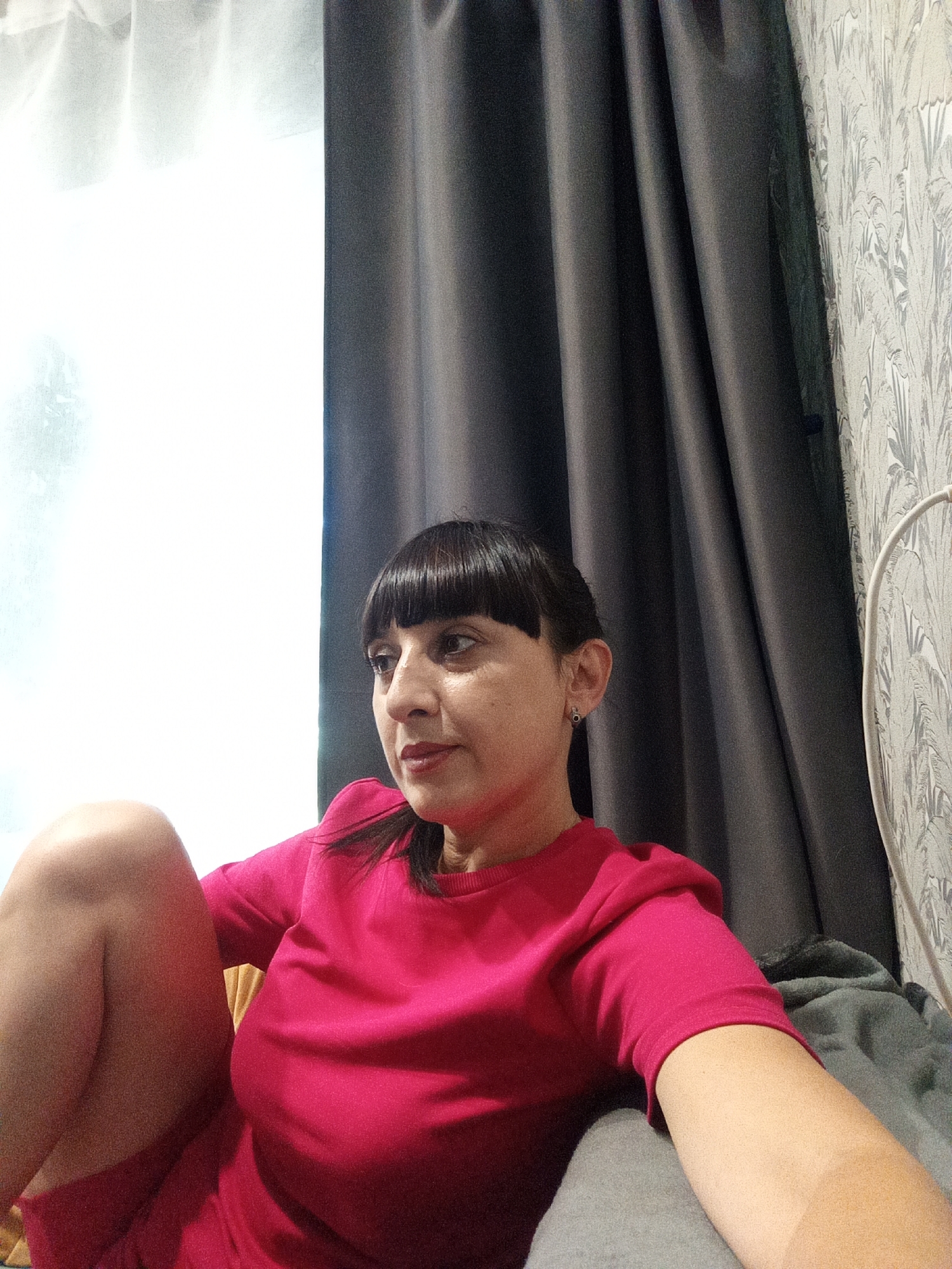 In Belogorsk, Russia, a woman with dark hair and bangs is captured from the chest up, seated indoors. She is wearing a vibrant pink top. Her gaze is directed to her left, with her head slightly tilted. The background features dark grey curtains to her right and a white curtain on the left side of the frame, suggesting a window is present. The lighting is bright, coming primarily from the left.