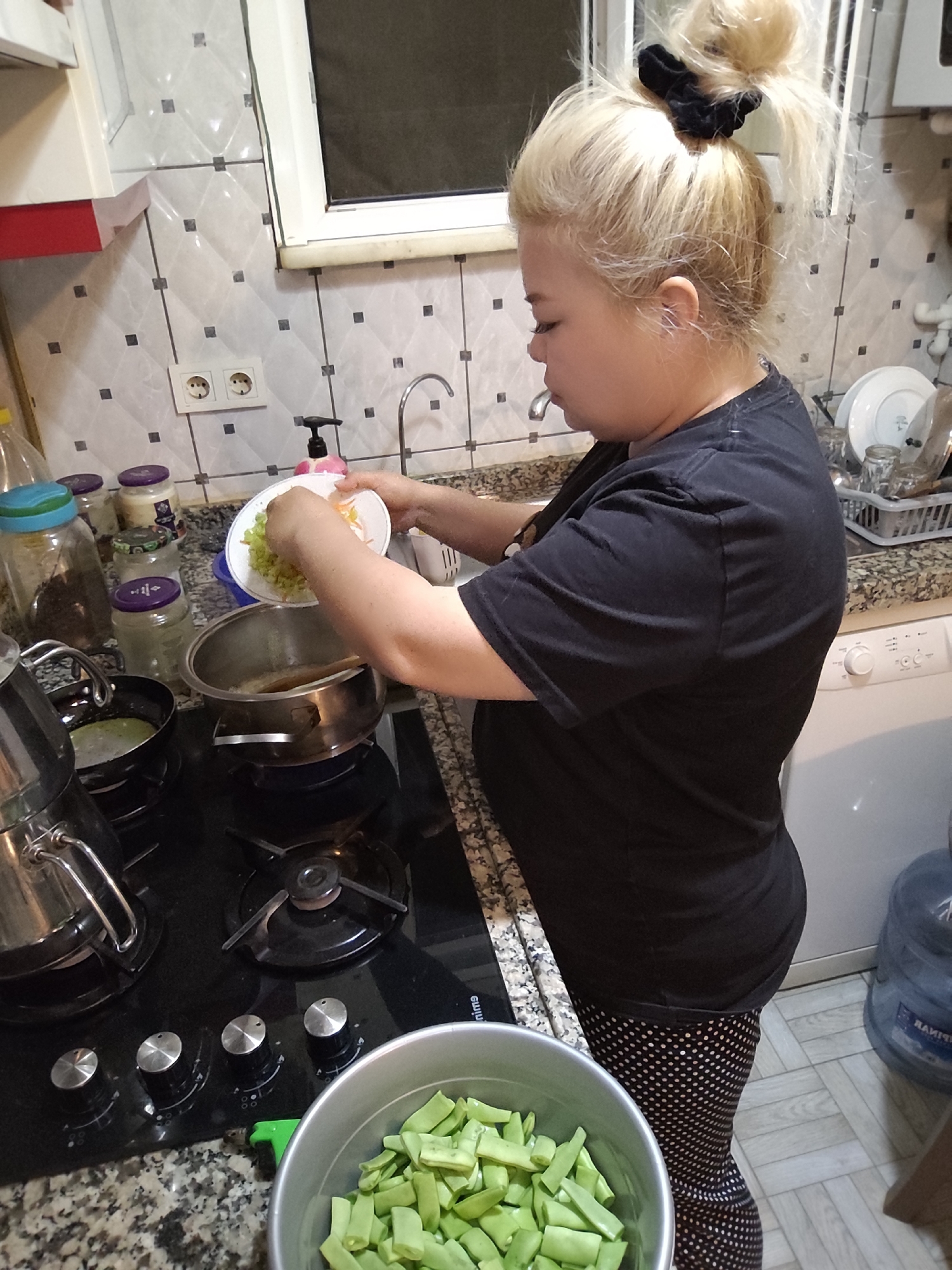 A woman is cooking in a kitchen in Sarıyer, Turkey. The woman has blonde hair in a bun and is wearing a black t-shirt and black and white polka dot pants. She is pouring the contents of a white plate into a stainless steel pot on a black glass stovetop. To the left of the stove is a kettle and several jars. In the lower foreground, there is a bowl of green beans. Behind the woman, there is a washing machine, a window, and a tiled wall.