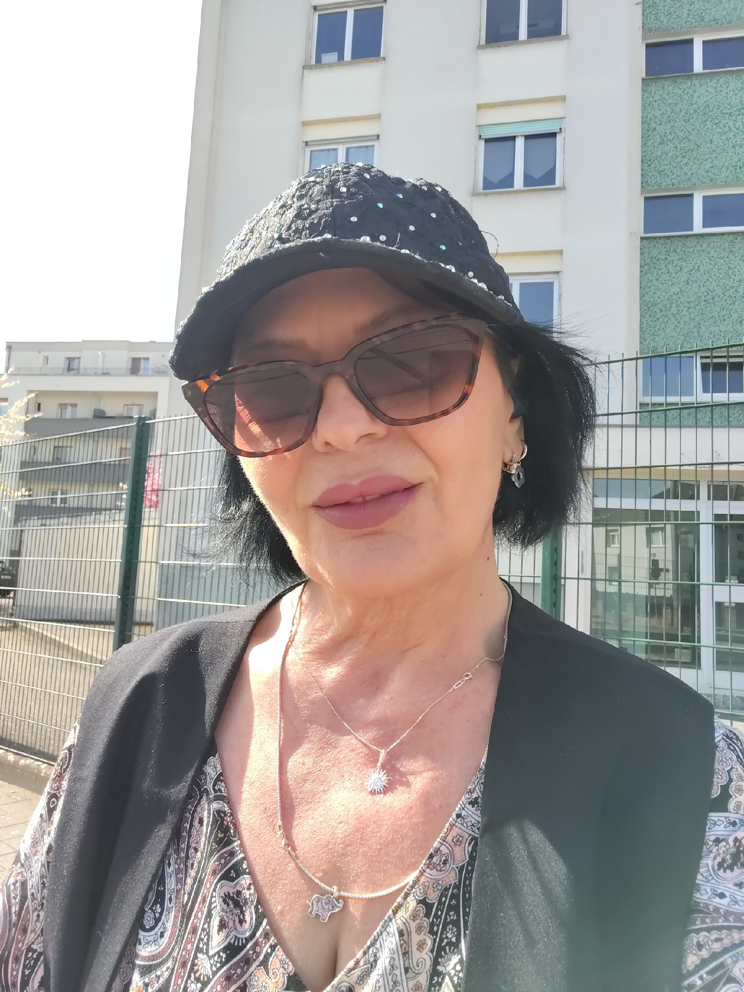A middle-aged woman with dark hair and sunglasses is taking a selfie outdoors. She is wearing a dark hat with decorative embellishments, a patterned top, and a black vest. Two necklaces are visible, one with a sun-shaped pendant and another with an elephant charm. She has earrings in her ears.
In the background, there is a multi-story building with windows and a fenced area. The location is possibly in Strasbourg, France, based on the architectural style of the buildings and the type of fencing. The lighting suggests it is daytime.