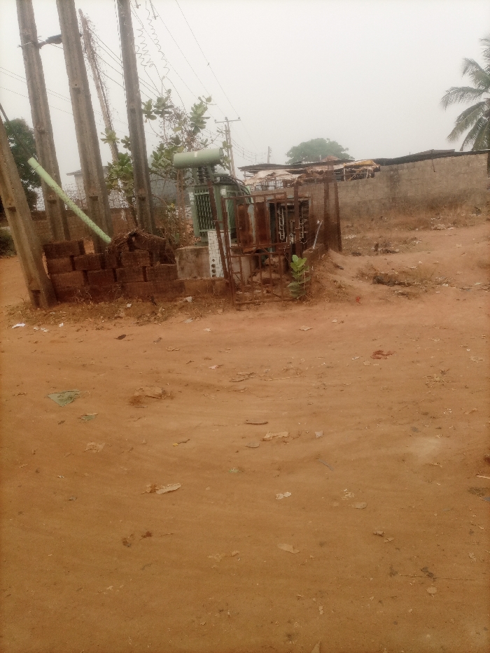 The image captures an outdoor scene, likely in a semi-rural or developing urban area, dominated by an electrical transformer station and utility infrastructure. The ground is unpaved, consisting of reddish-brown earth or sand, appearing somewhat uneven with small pieces of debris scattered across it.
In the center of the image, an electrical transformer is visible. It is a large, green, cylindrical unit mounted on a concrete base. Surrounding the transformer is a rusty metal fence or cage, indicating a lack of maintenance and possibly an older installation. Various electrical components, including insulators and wiring, are attached to the transformer and connected to overhead lines.
To the left of the transformer, multiple weathered utility poles, possibly made of concrete, stand tall, supporting a network of overhead power lines that stretch across the hazy sky. A light green pipe or conduit runs diagonally downwards from the left, seemingly attached to one of these poles, and partially disappears behind a stack of reddish-brown bricks or concrete blocks. These blocks appear somewhat broken and are haphazardly piled.
In the background, behind the transformer, there are simple building structures. One appears to be a grey concrete block wall, and other structures show signs of rudimentary construction, possibly with corrugated metal roofs. Scattered vegetation includes a small tree or large bush with green leaves near the transformer and a few smaller plants growing around the area. A palm tree is partially visible on the far right edge of the image.
The sky is overcast and hazy, creating a diffused, dull light across the entire scene. There are no strong shadows, suggesting either an overcast day or possibly atmospheric haze. No specific activity or event is taking place, and no text is visible in the image. The overall impression is one of an aged and somewhat neglected utility installation in an open, dusty environment.