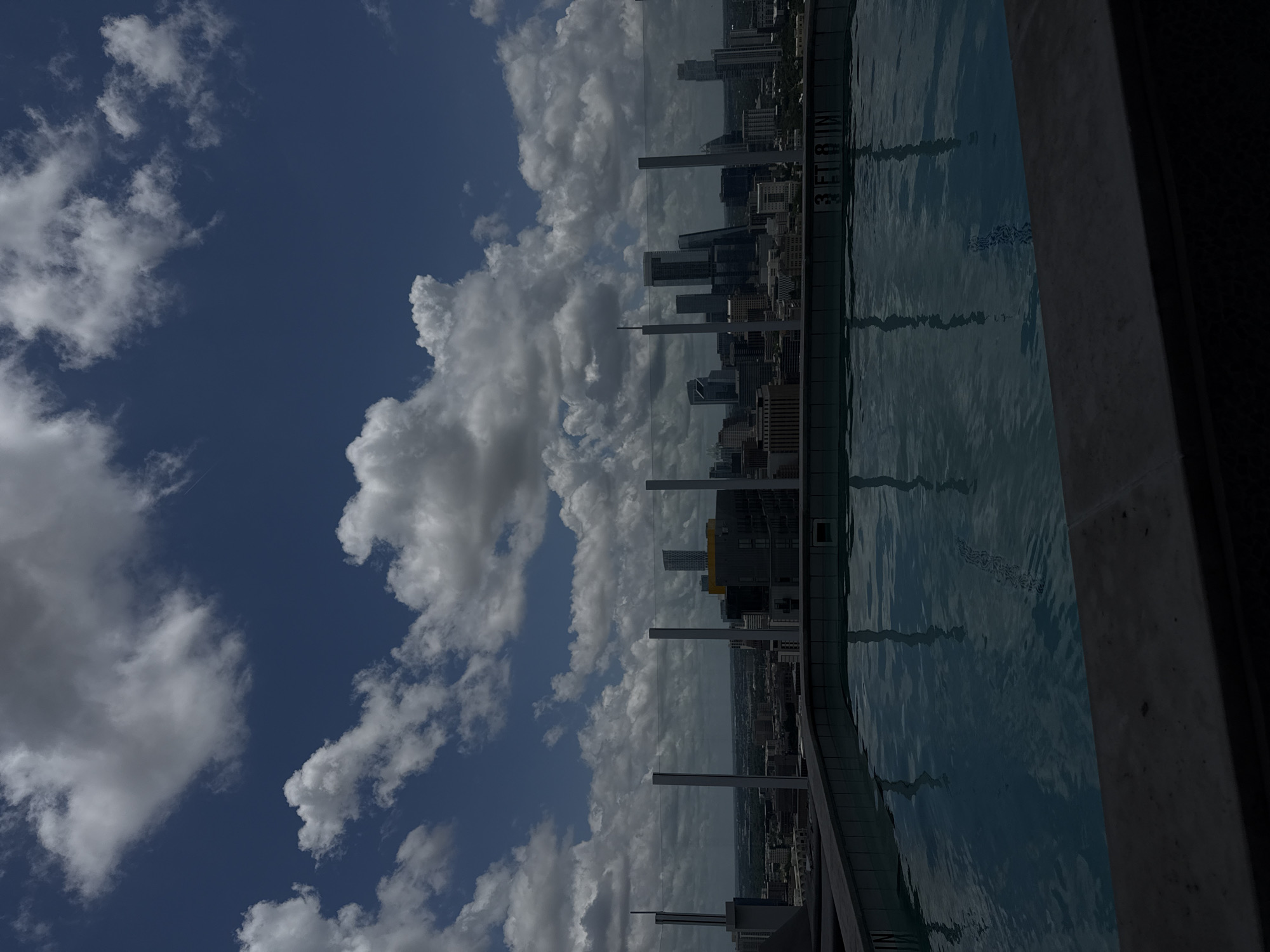 An elevated, static long shot captures a rooftop swimming pool overlooking the Austin, Texas, USA, downtown skyline under a partly cloudy sky. The foreground features the dark grey coping of the infinity-edge pool, with the pool's dark blue water occupying the lower portion of the frame. Reflections of the sky and distant buildings are subtly visible on the water's surface. On the far side of the pool, submerged white tiles reveal the depth marker "3 FT 8 IN" in black lettering. A transparent glass railing, supported by vertical metal posts, extends across the midground, separating the pool from the cityscape. Beyond the railing, the Austin skyline is prominent, featuring numerous high-rise buildings and skyscrapers under diffused daylight. Notable structures identifiable include the distinct 'Jenga'-like structure of The Independent on the right side of the main cluster, and the sleek design of the Austonian towards the center-left. The background consists of a deep blue sky dotted with numerous bright white and grey cumulus clouds, indicating partial cloud cover. No individuals are present in the scene, and no specific actions or interactions are depicted.