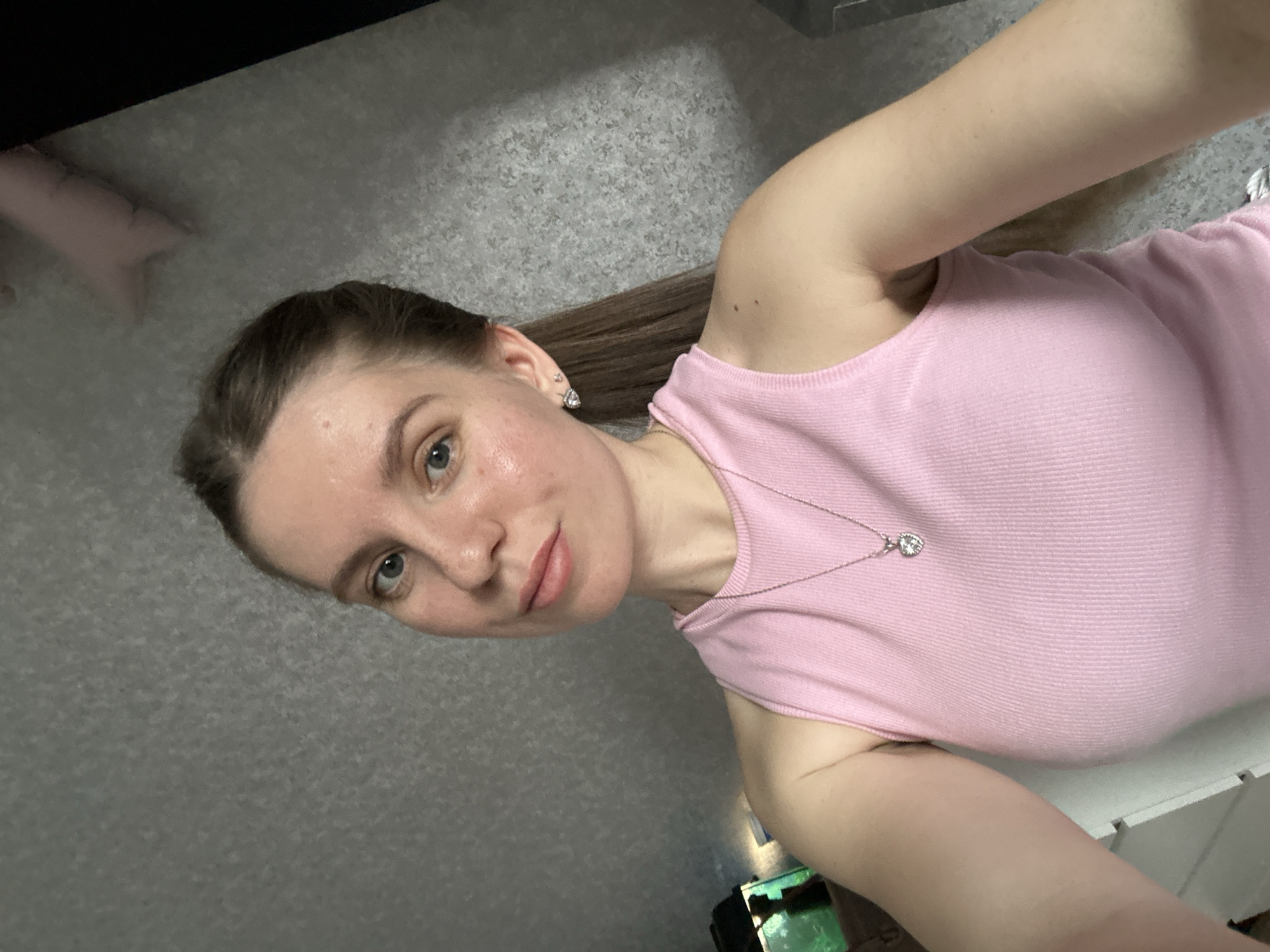 A woman with light skin and brown hair pulled back in a ponytail is positioned in front of a grey textured wall. She is wearing a pink tank top and a silver necklace with a heart-shaped pendant. Her face is slightly flushed, and she is looking directly at the camera. In the background, there's a glimpse of what appears to be furniture or a cabinet, and a small object with a green glow can be seen. There is no evidence to indicate the location is in Klin, Russia.