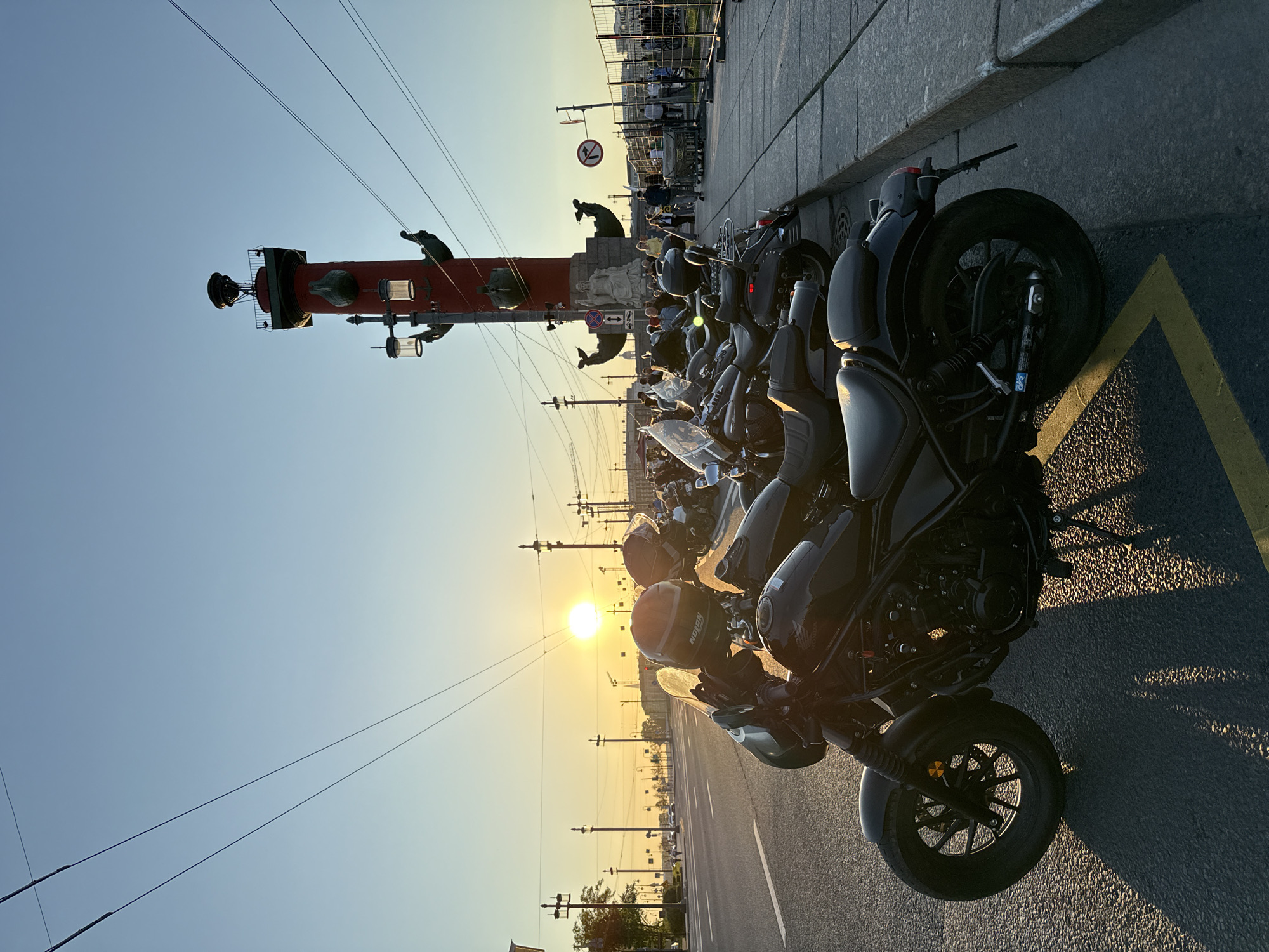 The image depicts an urban scene at sunset on Vasilyevsky Island in Saint Petersburg, Russia, featuring one of the city's Rostral Columns. In the foreground, a black Honda Rebel motorcycle is parked on asphalt, angled slightly to the left, with its side stand deployed. A black Nolan brand helmet rests on its handlebars. The motorcycle casts a long, dark shadow to the left. Behind it, a line of at least four additional motorcycles, predominantly cruiser-style, are parked along the curb. A yellow painted line is visible on the asphalt beneath the front wheel of the closest motorcycle.
To the right, a large section of a red Rostral Column is prominent, featuring sculpted ship prows and a white seated male statue at its base. The column extends upwards, crowned with an unlit brazier structure, partially intersected by overhead wires. A black street lamp post with two illuminated lanterns is affixed to the column. Several more street lamp posts line the wide asphalt road that extends into the distance on the left side of the frame.
The setting sun, a bright circular disk, is low on the horizon in the left-center, casting a strong golden light across the scene and illuminating the distant road. The sky transitions from a pale blue at the top to orange and yellow near the horizon. Multiple dark overhead power lines diagonally traverse the upper half of the image.
To the right of the Rostral Column, a paved pedestrian area is visible, occupied by numerous individuals. Metal barricades partially separate this area from an indistinct background. Various traffic signs, including a circular red sign with a diagonal bar indicating "no entry" or a specific prohibition, and a blue rectangular sign with an arrow, are mounted near the column.