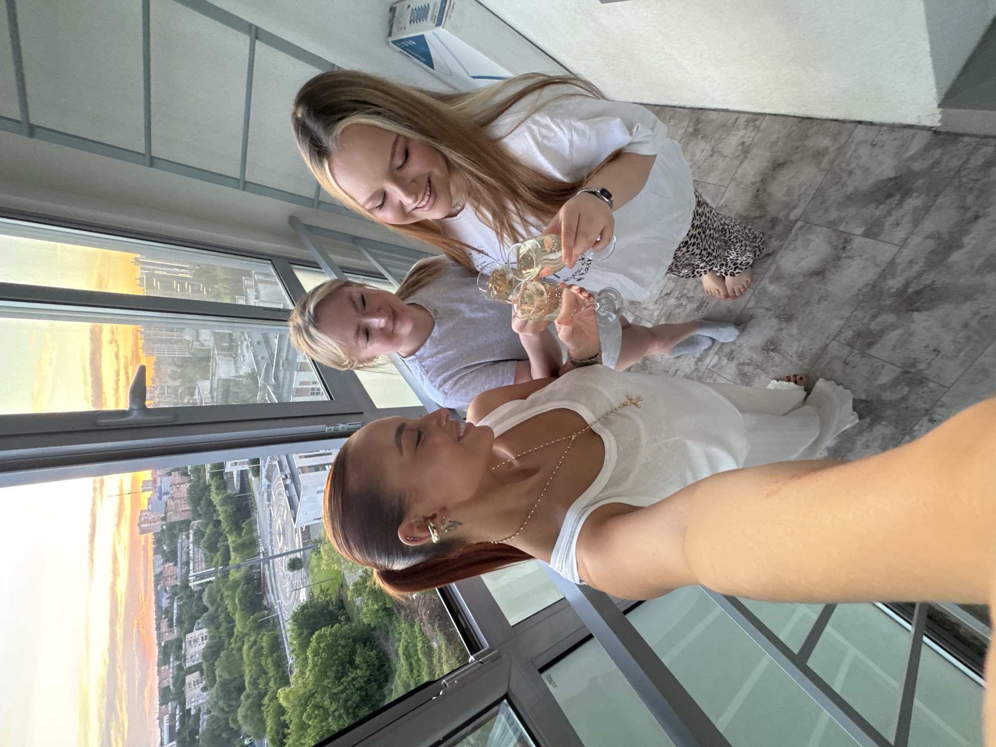 Three women are toasting with champagne in front of a window. The woman in the foreground has auburn hair pulled back into a ponytail, is wearing a white tank top, and has a gold necklace with a cross. The woman in the middle has blonde hair and is wearing a gray t-shirt. The woman in the background has long blonde hair with dark roots, is wearing a white t-shirt, and animal print pants. Outside the window is a view of a cityscape at sunset, with buildings and trees visible. The scene is in Izhevsk, Russia, as a roundabout is visible outside the window.
