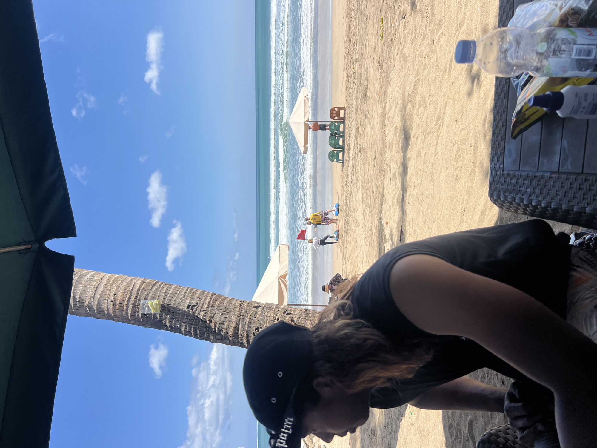 An outdoor view captures a beach scene in Kuta, Indonesia. In the foreground, a person wearing a black cap with "palm" printed on it and a black t-shirt sits on a wicker chair, partially obscured by the frame. The person's long, light-brown hair is visible. A dark green umbrella extends from the top left of the frame, providing shade. A palm tree trunk is prominent to the left of the person.
In the mid-ground, the sandy beach extends towards the ocean. Several beach umbrellas and lounge chairs are present, some occupied by people. Two red flags are planted in the sand near the water's edge. People are visible interacting in small groups.
The ocean in the background features moderate waves and a clear blue sky with scattered white clouds. A bottle of water and a bag of chips are placed on a table in the lower right corner of the foreground. The overall atmosphere suggests a sunny day at a tourist beach.