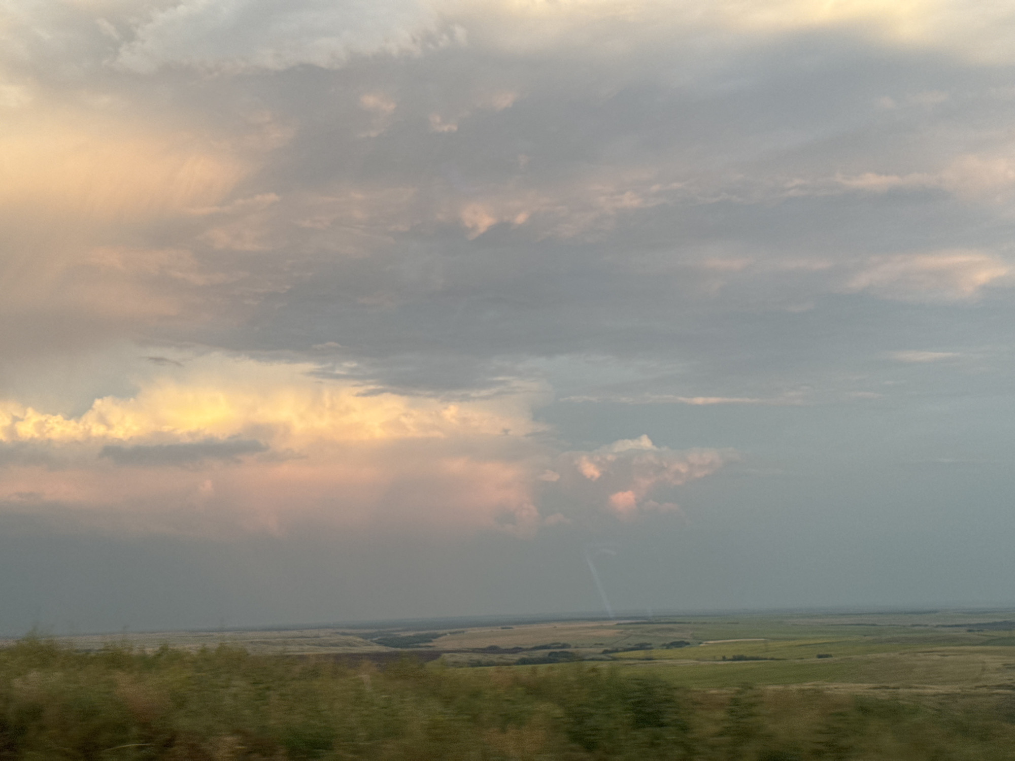 This is a wide shot of a vast, flat landscape under a dramatic sky. The horizon line is low, dividing the frame into a lower half of land and an upper half of sky. The land appears to be agricultural, with fields of varying shades of green and brown stretching out to the distant horizon. Some areas appear to be cultivated, with distinct patterns of crop rows, while others look more natural, with scrub and vegetation. The overall impression of the land is one of openness and expanse.
The sky is filled with a complex arrangement of clouds, suggesting either an approaching storm or the aftermath of one. In the left portion of the sky, the clouds are illuminated by warm, golden light, possibly from the sun setting or rising behind them, with soft pinkish hues mixed in. This warm light contrasts with the cooler, gray tones of the clouds that dominate the rest of the sky, creating a dynamic and moody atmosphere. A faint, vertical streak of light, possibly a wispy cloud or even a distant lightning strike, can be seen in the right mid-ground of the sky. The foreground of the image is blurred with streaks of green and brown, indicating movement, likely from the perspective of someone or something in transit. The overall lighting suggests it is either early morning or late evening. There are no people or identifiable man-made structures clearly visible in the scene. The scene depicts a natural, open environment with dynamic atmospheric conditions.