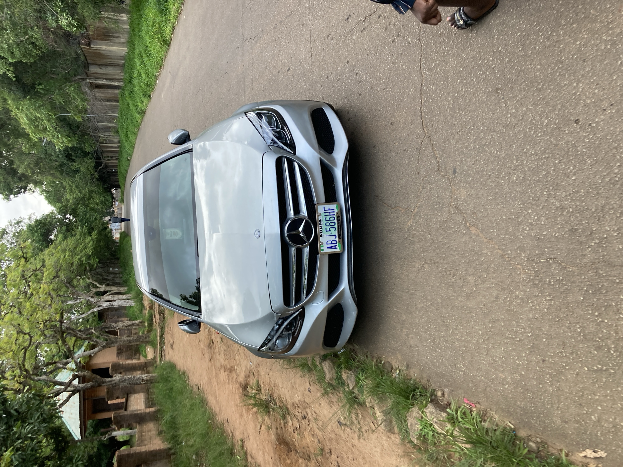 A silver Mercedes-Benz C-Class sedan is parked on a paved road, with its front facing the camera. The car's license plate reads "ABJ 586HF" and indicates "Abuja" and "Mercedes-Benz." The car is positioned slightly to the right of the road's center. To the left of the car is a dirt path bordered by overgrown grass and a low brick wall. Beyond the wall, a single-story building with a corrugated metal roof is visible, surrounded by trees. The background features a row of trees lining the right side of the road, with a concrete wall behind them. A person is walking away from the camera in the distance on the left side of the road. The right side of the frame shows a person's legs and feet, wearing sandals, standing near the edge of the road. The environment appears to be a suburban or semi-rural area, possibly in the city of Jos, Nigeria, given the context provided by the license plate and the architectural style of the buildings. The lighting suggests it is daytime, with overcast skies.