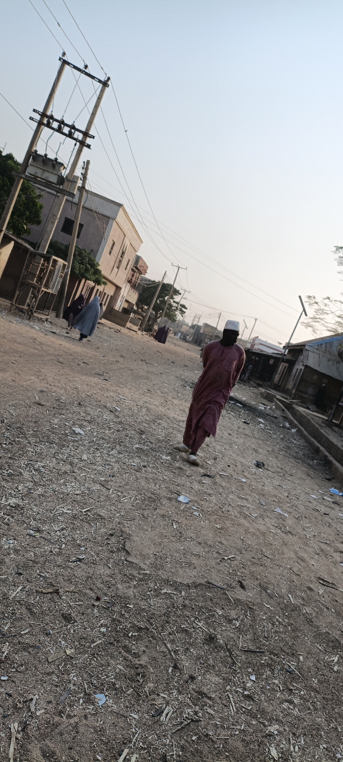 A street scene shows a man wearing a maroon robe and white cap walking towards the viewer. Another person in a full blue robe walks in the opposite direction. The unpaved street is filled with debris. Buildings line both sides of the road. Power lines are visible above. The sky is a light blue.