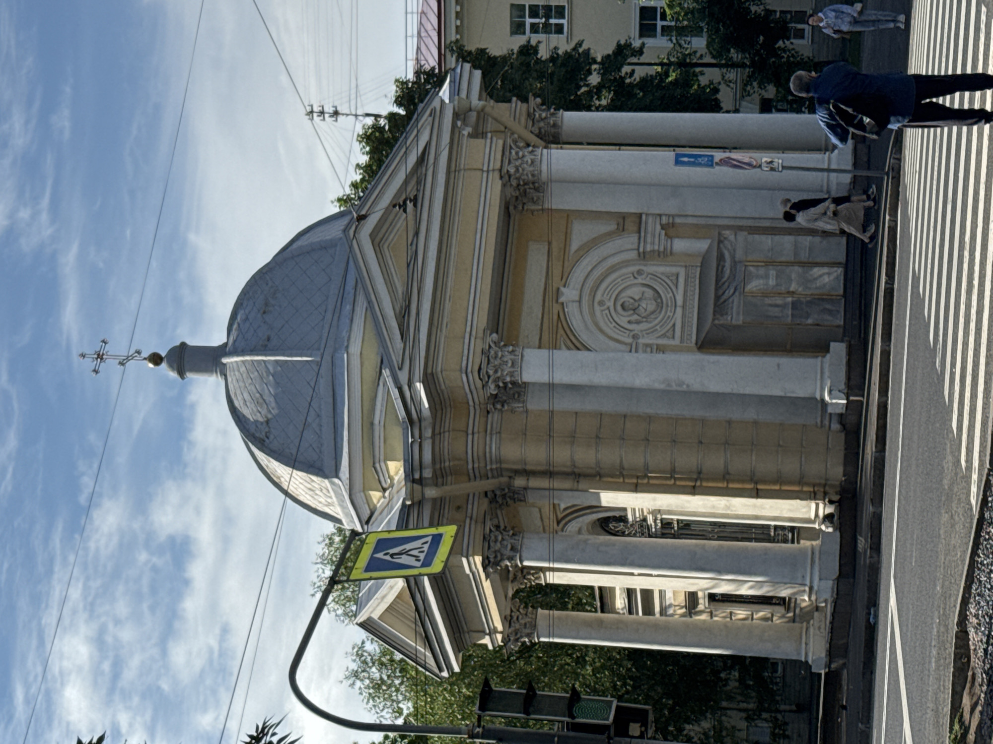 A yellow, neoclassical building with a dome topped by a cross stands at a street intersection in Saint Petersburg, Russia. The building has white columns and decorative reliefs. A pedestrian crossing is visible in the foreground. A traffic light with a green light illuminated hangs from a pole on the left. A pedestrian crossing sign is also visible hanging from a pole. Several people are walking on the sidewalk near the building, and one person is standing at the crosswalk with their back to the camera, wearing a blue jacket and backpack. The sky is blue with scattered white clouds. The scene appears to be captured in daylight.