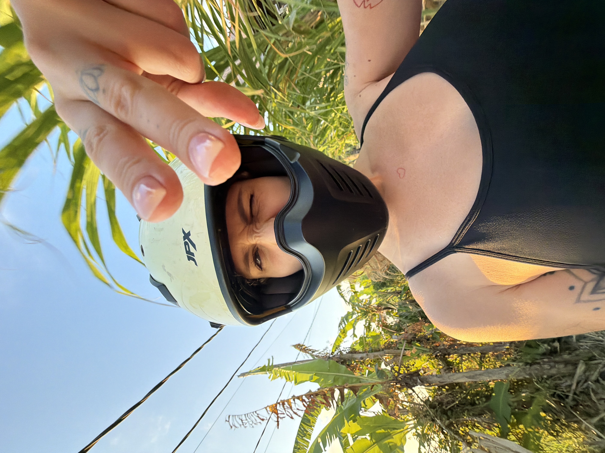 An outdoor, eye-level, medium shot captures a person from the chest up, wearing a light-colored motorcycle helmet with a black chin guard and a black tank top. The person is positioned slightly off-center to the right and is looking at the camera with one eye closed, the other squinting. The person's right hand is extended towards the camera, with fingers partially obscuring the helmet. The background features lush green foliage, including banana trees, and power lines overhead against a bright blue sky. The lighting suggests it is late afternoon or early morning. Based on the vegetation and general atmosphere, the location is consistent with the tropical environment of North Kuta, Indonesia.