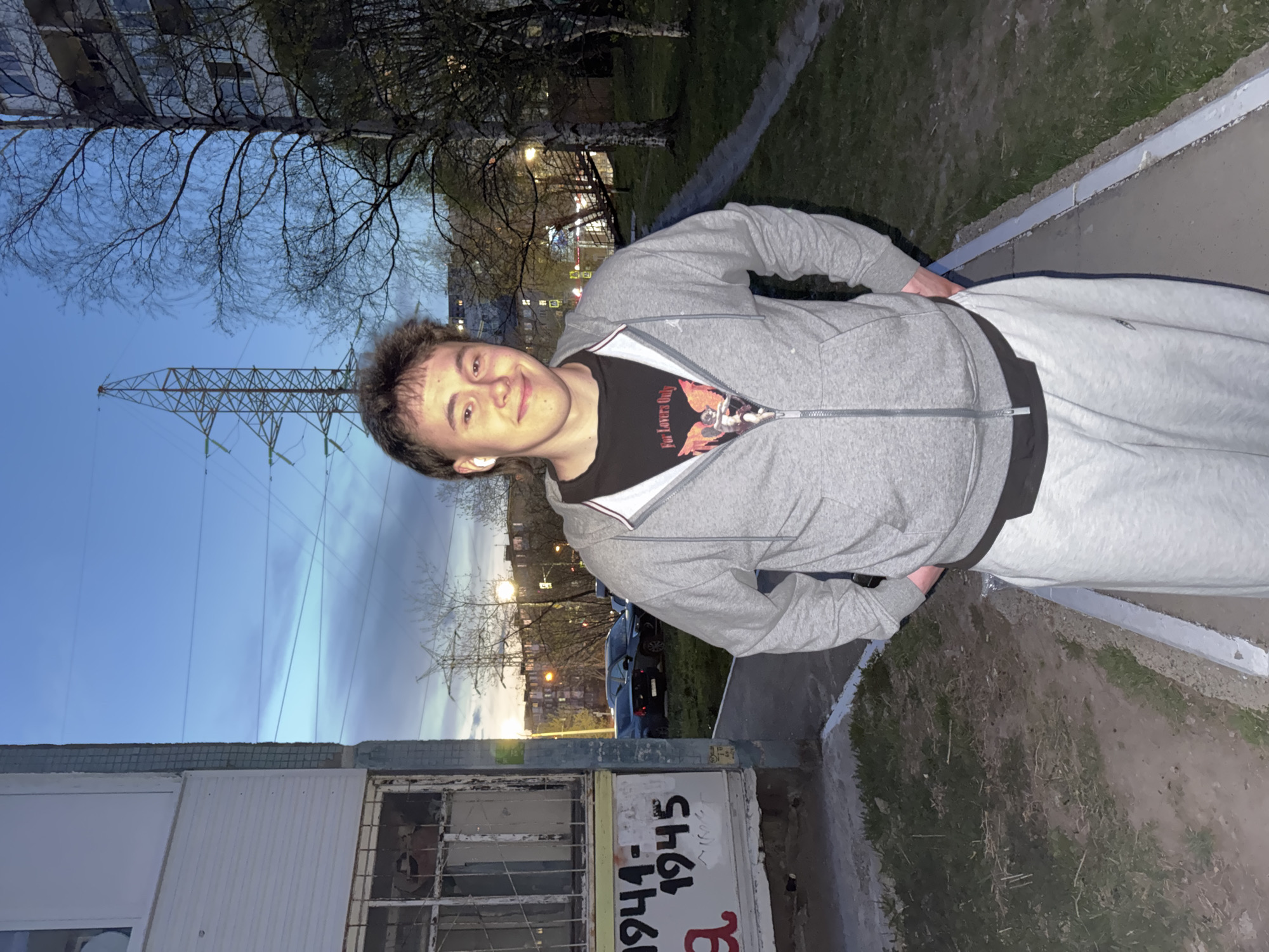 A young man is standing outdoors near a building. He is wearing a grey zip-up hoodie over a black t-shirt and light grey sweatpants, and appears to have an earpiece in his left ear. He is smiling slightly. In the background, an electrical transmission tower is visible against a dusk sky. The building next to the man is marked with the numbers "1944-1945". The location could be Perm, Russia.