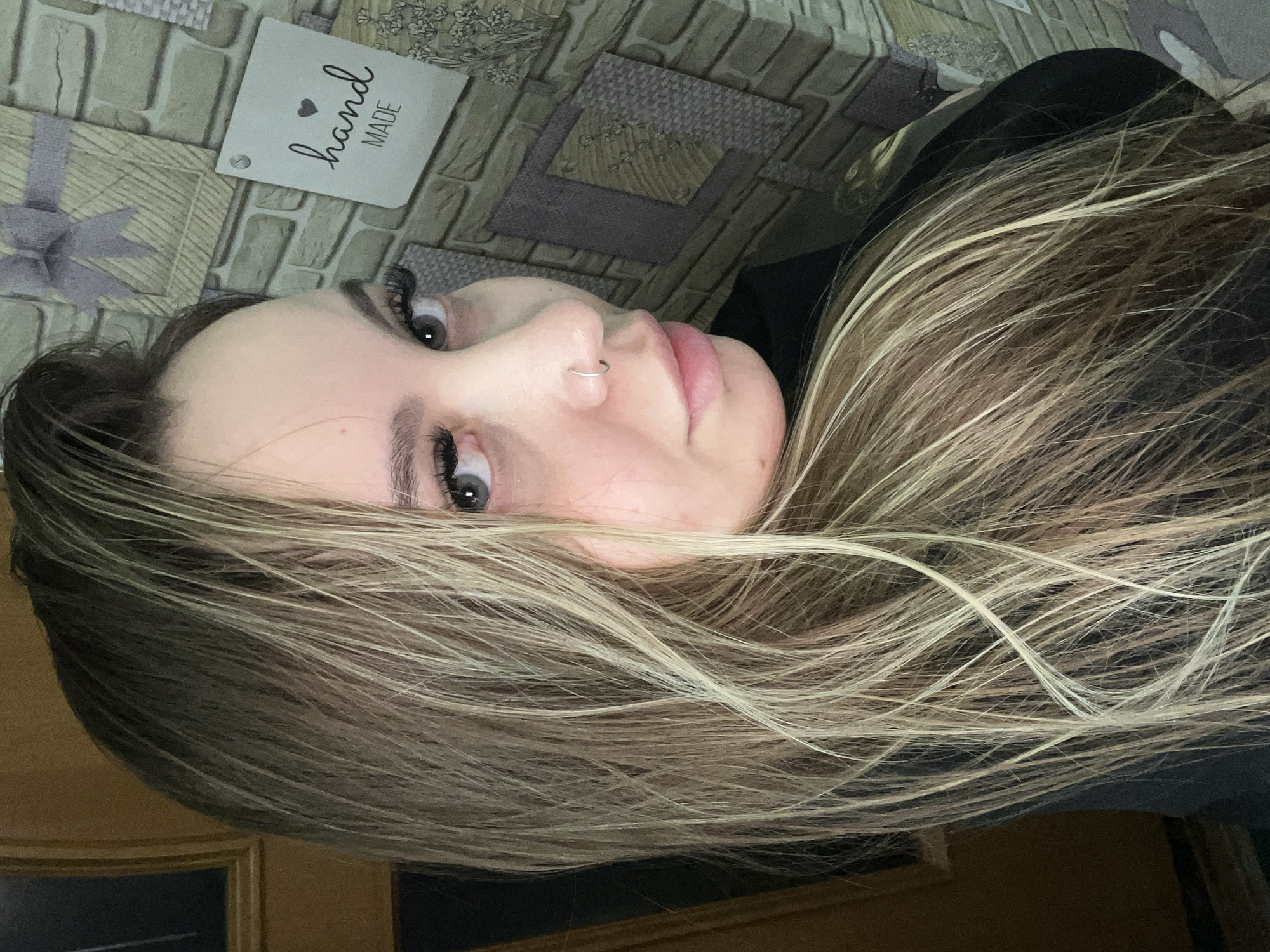 A close-up, tilted view of a light-skinned woman with long, highlighted brown hair. She has gray eyes and a nose ring. She is wearing false eyelashes. She is lying on a bed with a patterned bedspread. In the upper left corner is a tag that says "hand MADE" with a heart above the word "hand". The bottom of a door is visible in the lower part of the image. There is no information to indicate this image was taken in Lyubimov, Russia.