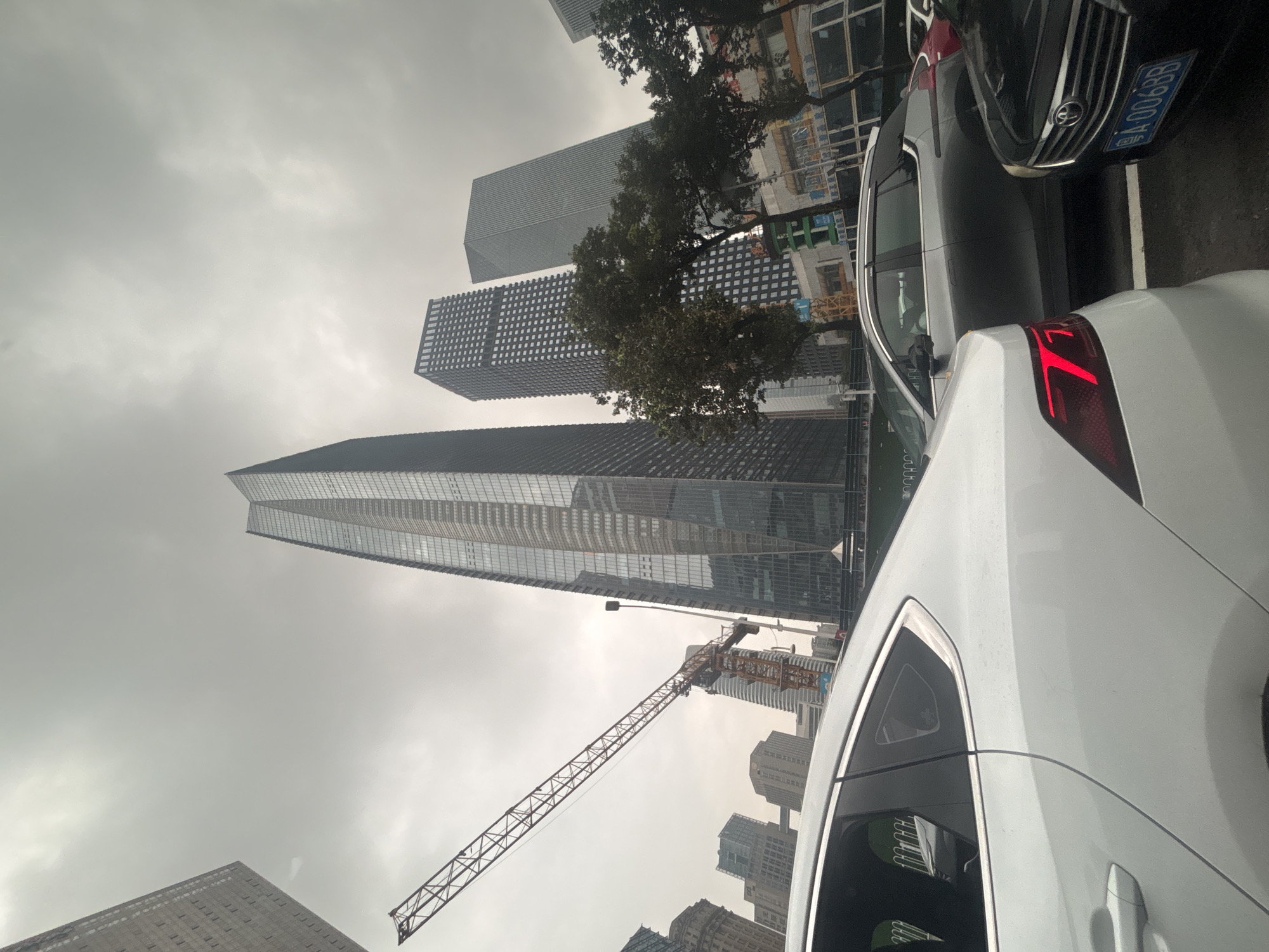 This is a low-angle, wide shot of a city street in Tianhe District, China, on an overcast day. In the foreground, a white sedan is parked, its rear quarter visible. To its right, a black sedan is also parked. Further down the street, a grey SUV is visible. Dominating the background are several modern skyscrapers, the tallest of which is a glass-fronted building with a unique angular design. To the left of the skyscrapers, a construction crane extends diagonally across the sky. The sky is heavily clouded and appears grey. Trees line the street between the vehicles and the buildings. License plates are partially visible on the black and grey vehicles. The overall impression is one of an urban environment with ongoing construction.