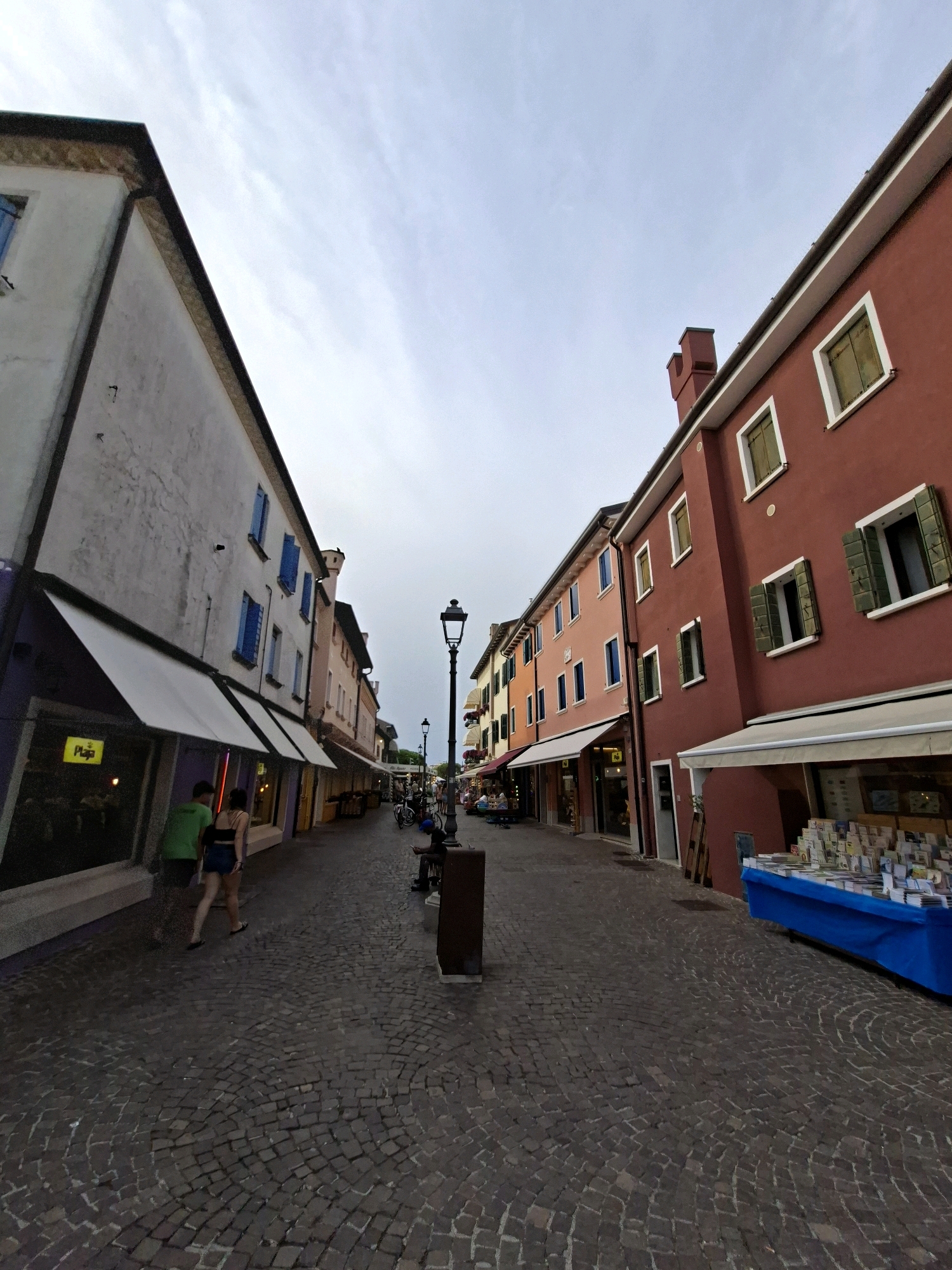 The image depicts a street scene in Caorle, Italy, characterized by its narrow cobblestone alleyway lined with buildings of varying colors. On the left, a white building with a purple storefront is visible, featuring awnings and a yellow "Plaja" sign. Two individuals, a man in a green t-shirt and a woman in a black top and denim shorts, walk away from the camera on the left side of the street.
The right side of the street features buildings painted in shades of red, orange, and pink. The red building, prominently in the foreground on the right, has multiple windows with green shutters. A blue-covered table displaying books or other merchandise is set up outside this building. A lamppost stands in the middle of the street, and several bicycles are parked further down. The sky above is overcast. The paving stones of the street are arranged in a circular pattern.