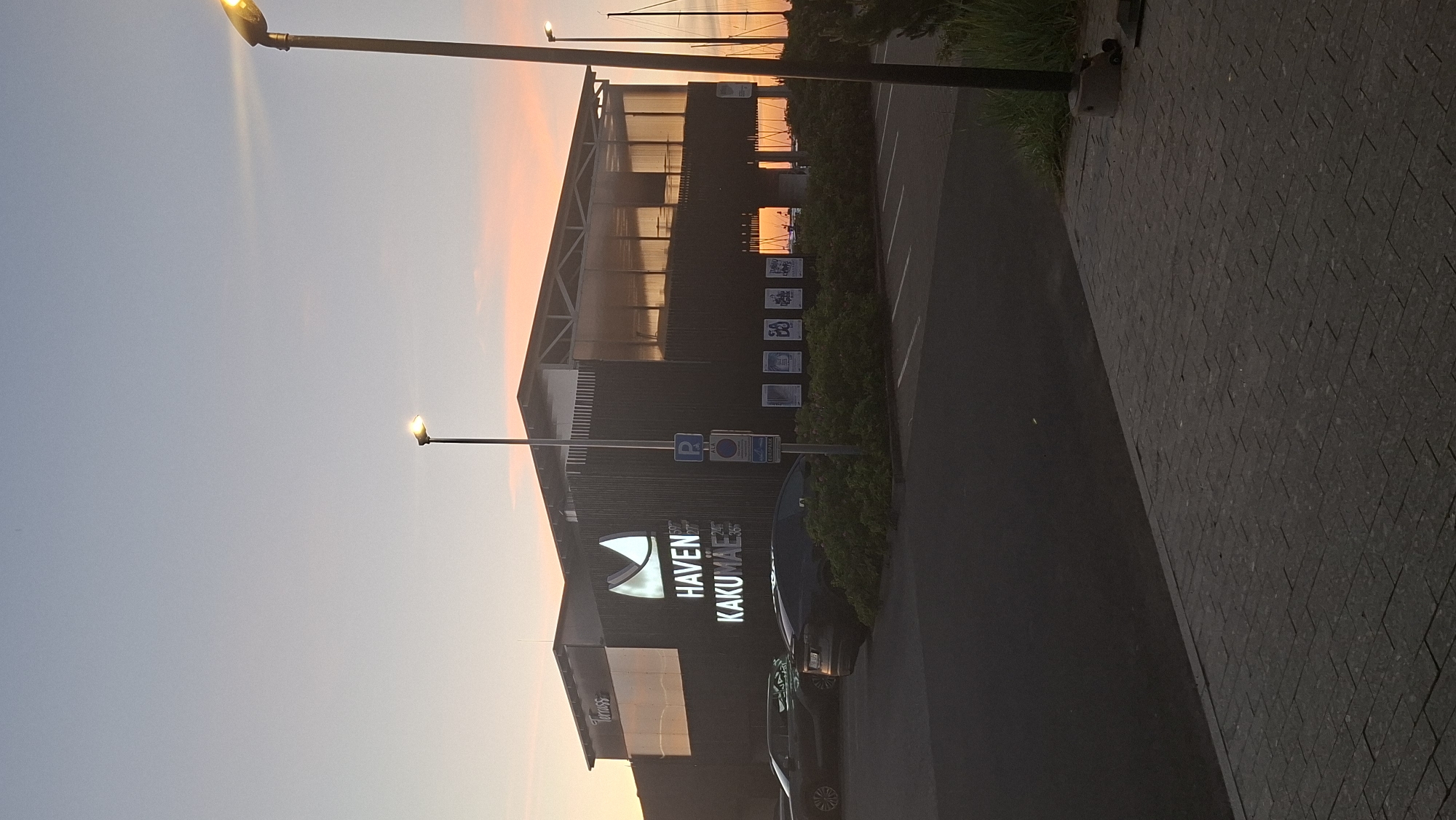 An exterior, medium shot shows a modern commercial building at sunset, likely a restaurant or entertainment venue, named "HAVEN KAKUMÄE." The building features dark vertical siding and a glass-enclosed upper level that reflects the colorful sky. Two cars are parked in front of the building, a dark sedan on the left and a black SUV on the right. A streetlamp with an illuminated bulb stands to the right of the building, and another streetlamp is visible in the distance. The foreground is a paved walkway with a dark asphalt parking lot extending behind it. The sky is a gradient of pale blue at the top to orange and pink near the horizon, indicating it is either sunrise or sunset. The overall scene is peaceful and serene.
