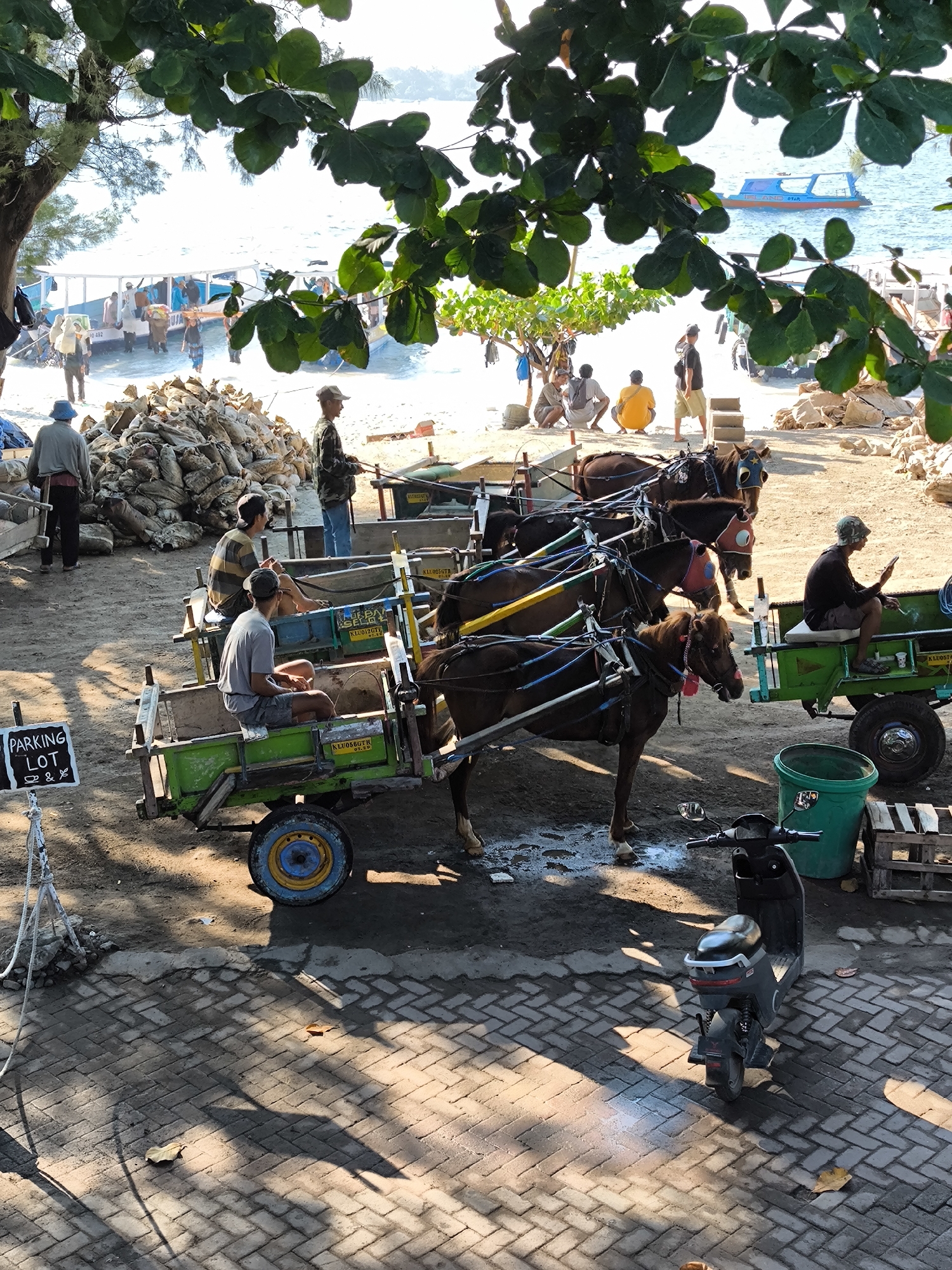 The image depicts a scene in an unspecified location in Unknown, Indonesia, featuring a row of horse-drawn carts lined up on a brick-paved area. Four carts are visible, each pulled by a brown horse. The drivers sit in the carts, with one occupied and three not. A sign reading "Parking Lot" is positioned to the side. A scooter is parked nearby. In the background, people are gathered near a pile of sacks on the beach. The ocean and a boat are visible in the distance. Green foliage frames the top of the image.