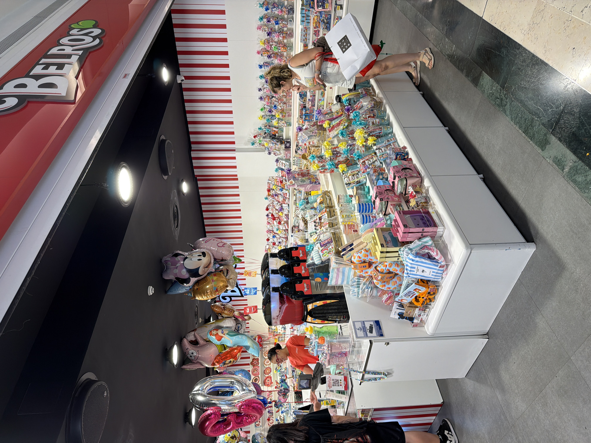 An interior shot captures a brightly lit candy store called "BELROS" located in an unknown city. The store features a white counter with various candy displays and a slushie machine. A female employee in an orange shirt stands behind the counter, interacting with a customer, a female with dark hair wearing a black t-shirt. In the foreground, a woman with blonde hair, wearing a white top and shorts, examines a gift bag from "RITUALS." The store's interior is decorated with red and white striped walls and is adorned with numerous balloons, including character balloons like Mickey Mouse and Patrick Star. The floor is a mosaic of grey and green tiles.