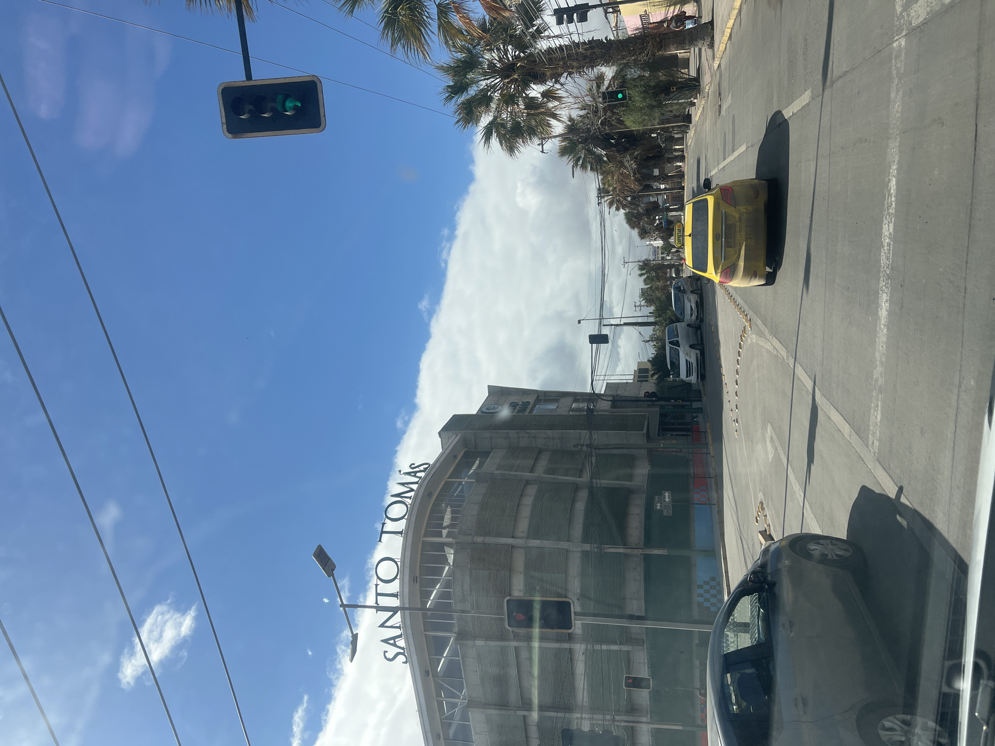 The image shows a street scene in Arica, Chile, with the building of "SANTO TOMÁS" prominently visible on the left. The sky is blue with scattered white clouds. There are several cars on the road, including a silver sedan on the left and a yellow taxi in the center lane, moving away from the viewer. Traffic lights are visible; one is green above the yellow taxi, and another red light is visible on the building's side. Palm trees line the right side of the street. The road has clear lane markings and crosswalks. The building appears to be under construction or renovation, with some scaffolding visible. The overall impression is a sunny day in an urban environment.