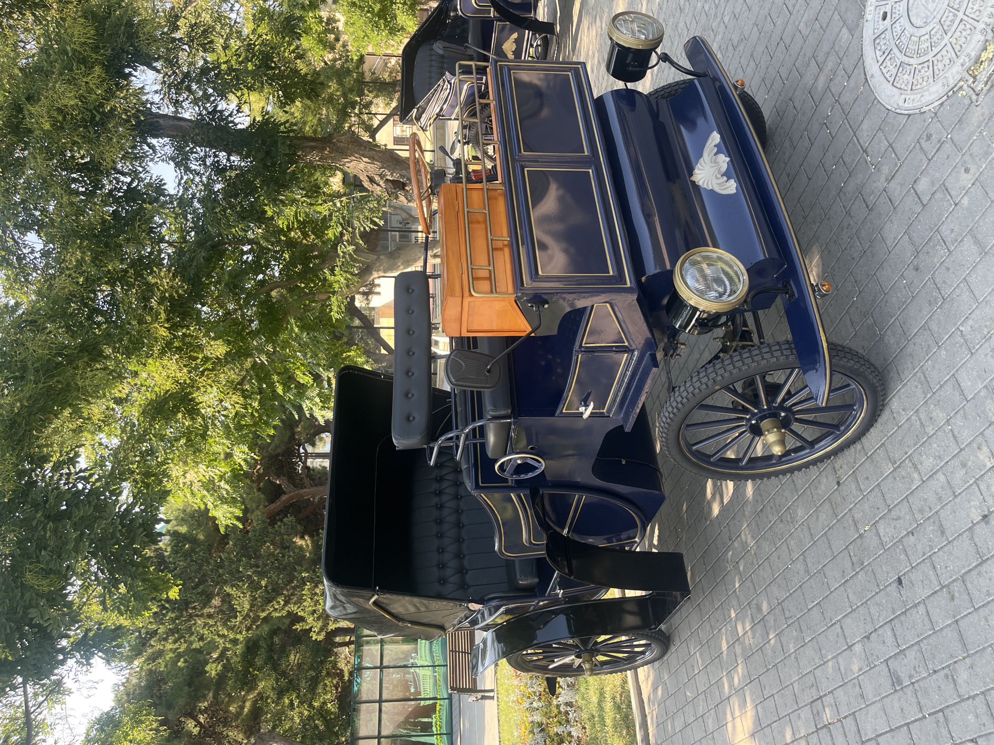 A navy blue antique car with black upholstery and gold trim is parked on a paved walkway. The car has two headlights on the front, a wooden box on the rear, and a black canvas roof. In the background, there are trees, a bench, and a building with a glass facade. The image is taken in Baku, Azerbaijan.