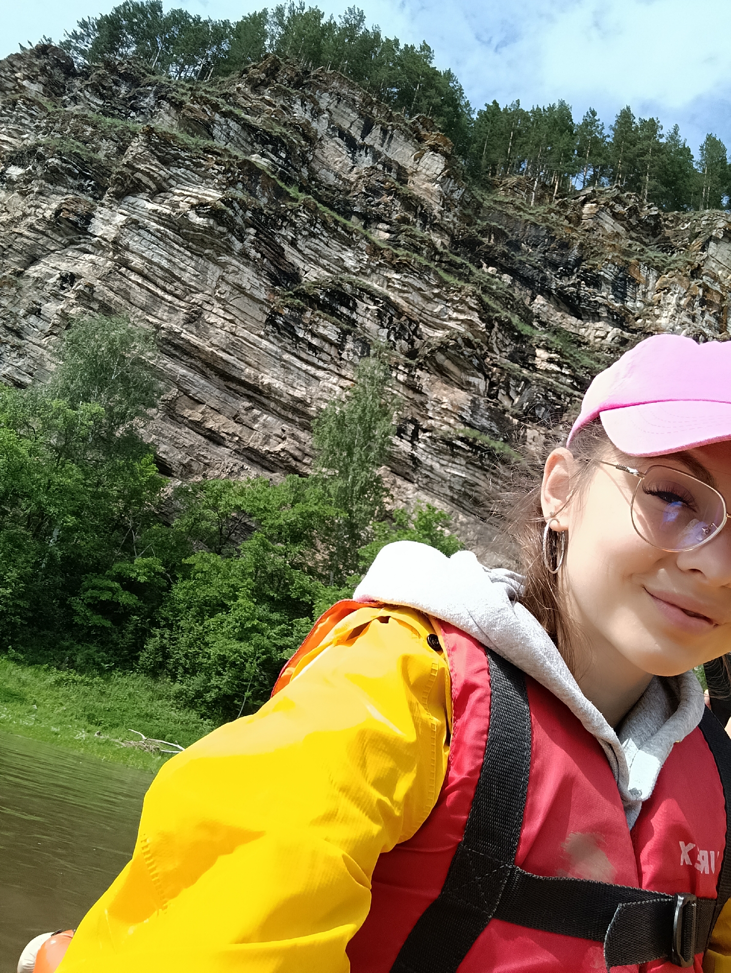A young woman in a pink baseball cap and glasses is captured in a selfie, smiling. She is wearing a yellow jacket over a grey hoodie and a red life vest, suggesting an outdoor activity such as rafting. The background features a steep, layered rock cliff face covered in sparse green vegetation and trees. Below the cliff, a body of water, likely a river, is visible. The sky is partly cloudy. The image appears to have been taken during daylight. No specific location within Sergeevka, Russia is identifiable.