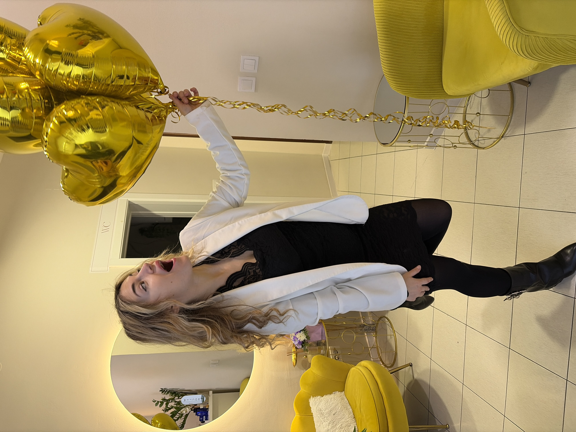 A woman stands indoors, holding a bunch of gold heart-shaped balloons with attached gold ribbons. She is wearing a black dress, white blazer, black leggings, and black boots. Her mouth is open in what appears to be excitement. The setting seems to be a decorated room featuring yellow upholstered chairs and a gold-colored round mirror on the wall. A sign that reads "WC" is visible above a doorway. The exact location in Budva, Montenegro, cannot be determined from the image alone.