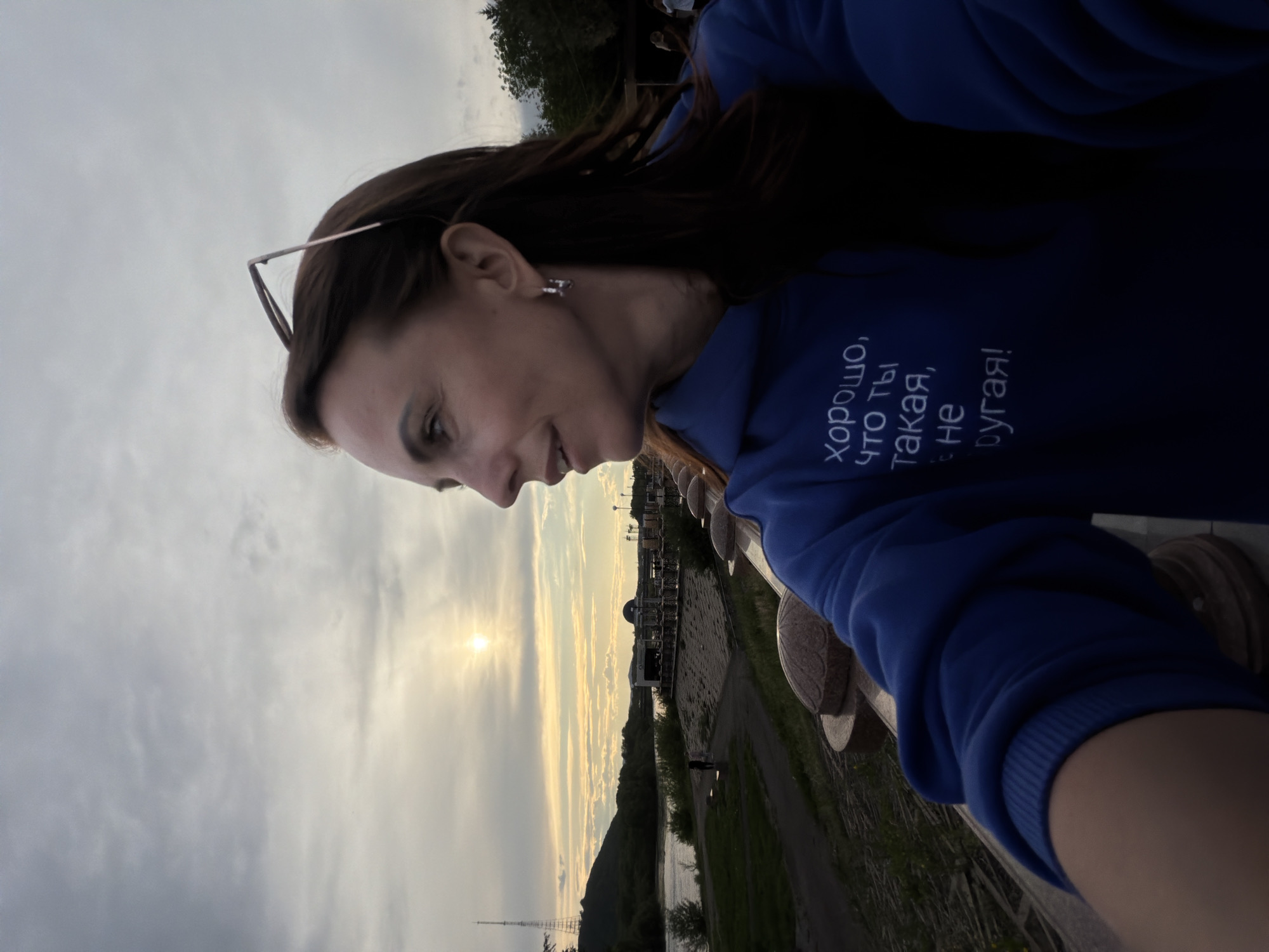 A woman is captured in a selfie, positioned outdoors against a scenic backdrop, likely in Birobidzhan, Russia. She is shown in profile, smiling gently, with dark hair and a silver earring visible. Her glasses are pushed up onto her head. She wears a dark blue hoodie featuring white Cyrillic text that reads: "Хорошо, что ты такая, не другая!" This translates to "It's good that you are like that, not another (different) one!"
The woman appears to be standing on a bridge or promenade, with a wooden railing partially visible in the foreground. Beyond her, a body of water stretches across the middle ground, bordered by land with trees and distant buildings or structures. A tall mast or antenna is prominently visible on the left side of the distant landscape. The sky is partly cloudy, with the sun low on the horizon, indicating either a late afternoon sunset or early morning sunrise, casting a soft, warm light over the scene. The overall mood conveyed is peaceful and happy.