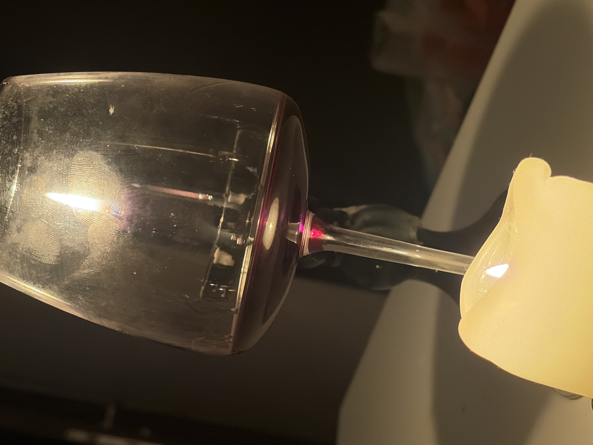 A red wine glass, containing a small amount of red wine, rests horizontally, partially obscuring a lit, off-white candle. The wine glass is dirty, bearing visible fingerprints. The background is dark and blurred, suggesting an indoor environment. The scene is not identifiable to a specific location in Budva, Montenegro, based on the visual information.