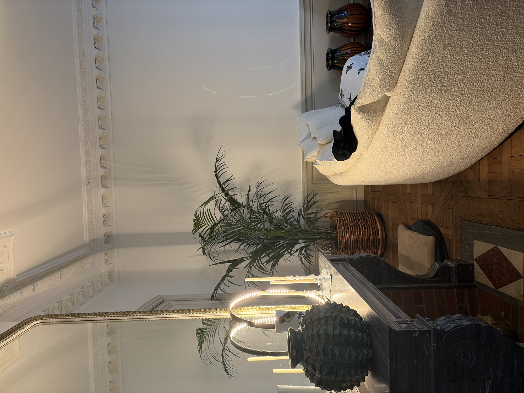 This image depicts a finely appointed interior room, likely an apartment in Paris, France, given the distinctive Haussmannian architectural elements such as the ornate ceiling mouldings, high ceilings, classical fireplace, and parquet flooring.
The scene features a fireplace mantel on the left, constructed from dark marble or stone, with a large, heavily textured, dark green or bronze urn-like object positioned prominently on its surface. To the right of the urn, several white tapered candles are lit in various holders, emitting warm light. Behind these candles, a circular LED ring light illuminates a small decorative object, possibly a vase with a bird motif, and reflects in a tall, gold-framed mirror that extends upwards, reflecting parts of the room, including a large potted plant.
To the right of the fireplace, a substantial green palm plant sits in a woven basket on the wooden floor. Further to the right, occupying a significant portion of the frame, is a large, curvilinear sofa upholstered in a light-colored, textured fabric, possibly bouclé. White throw pillows are arranged on the sofa, with a dark garment draped over the back and a patterned blanket partially visible.
The floor is made of wood with a patterned parquet design. Directly in front of the fireplace, a tiled hearth displays red and white diamond patterns. A small, beige pet bed or cushion rests on the floor next to the fireplace's base. Behind the sofa, against the wall, two large ceramic vases with brown and orange vertical stripes are visible. The walls are painted white above a grey wainscoting, and the ceiling features intricate white plaster cornices and mouldings. The overall lighting is dim and ambient, emanating primarily from the candles and ring light, creating warm reflections and soft shadows throughout the room. No individuals are present, and no overt actions are occurring.