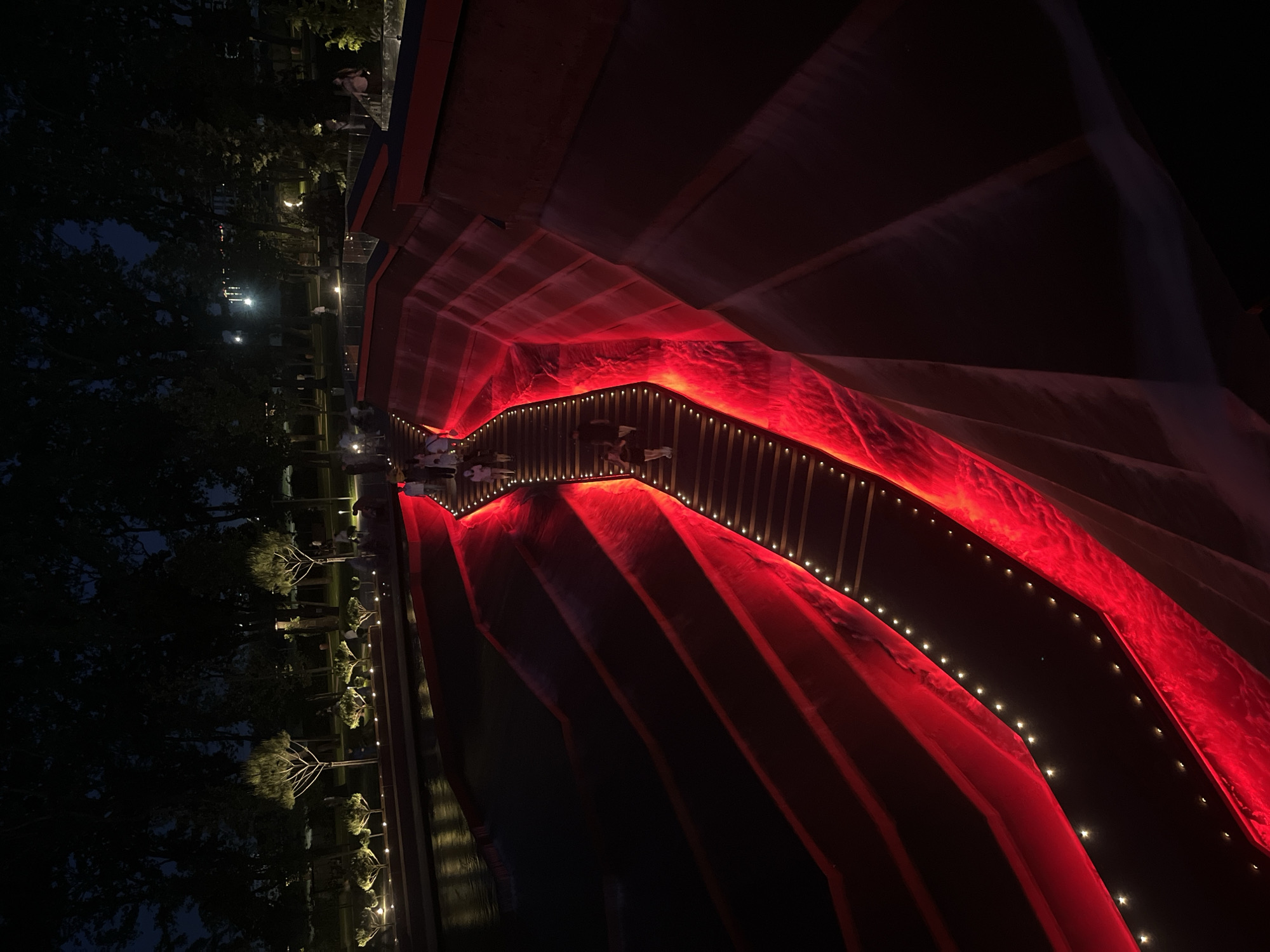 The image shows an illuminated outdoor staircase at night. The staircase is carved into a tiered, reddish-brown structure and is lit from below with bright red lights, creating a dramatic effect. Small, warm-toned lights are also embedded along the edges of the stairs. Several people are walking up and down the staircase. In the background, trees are silhouetted against a dark sky, and a body of water reflects some of the ambient light. The scene appears to be a public space, likely a park or promenade. This location is the "Oasis" park in the city of Krasnodar, Russia.