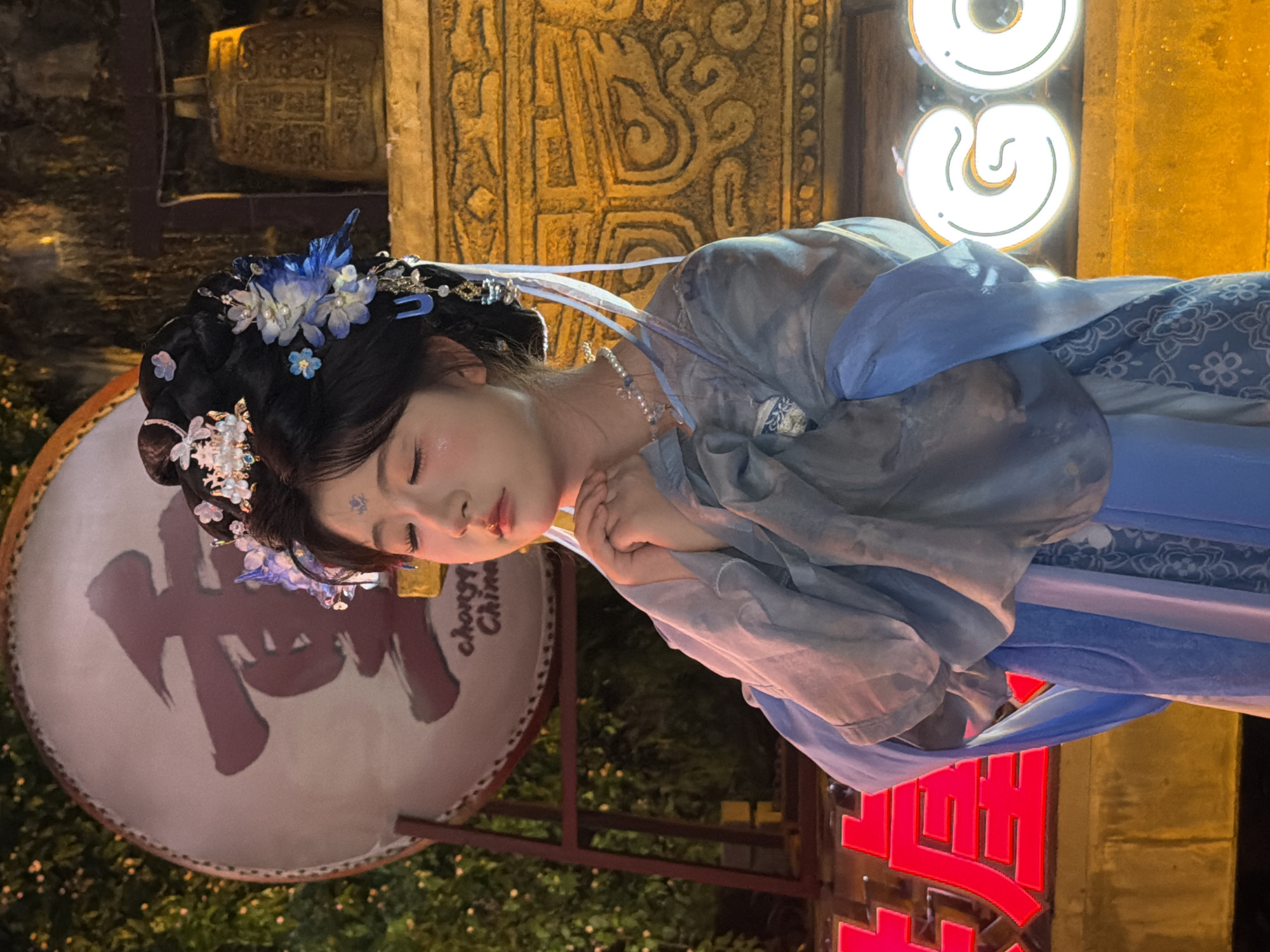 A young woman dressed in traditional Chinese attire, possibly a hanfu, is depicted in this image. She has black hair adorned with floral accessories and delicate hairpins, and her face is softly illuminated, suggesting a close-up or portrait shot. Her eyes are closed, and her hands are clasped in front of her chest, conveying a sense of peace or contemplation.
The woman is positioned in front of a large, circular object that appears to be a drum, on which a bold, dark Chinese character is painted. To the left of the woman, there is an ornate, golden-toned architectural element with intricate carvings. In the background, there are bright, glowing circular lights, possibly part of a sign or decoration, and a neon sign with bold red Chinese characters is visible in the lower right portion of the frame. The overall scene is illuminated by warm, artificial lighting, indicating that it is likely nighttime or a dimly lit indoor setting. The woman's makeup includes a subtle blue design on her forehead. The dress she is wearing is layered, with flowing sleeves and a blue sash.
The visible text includes Chinese characters on the drum, a partial inscription that reads "chorgn China" on the drum as well, and red Chinese characters on the neon sign. The presence of traditional attire, architecture, and Chinese characters suggests a cultural setting, possibly a festival, performance, or a tourist attraction in China, specifically noted as Yuzhong District. The mood of the image is serene and elegant, focusing on the beauty of the subject and the cultural context.