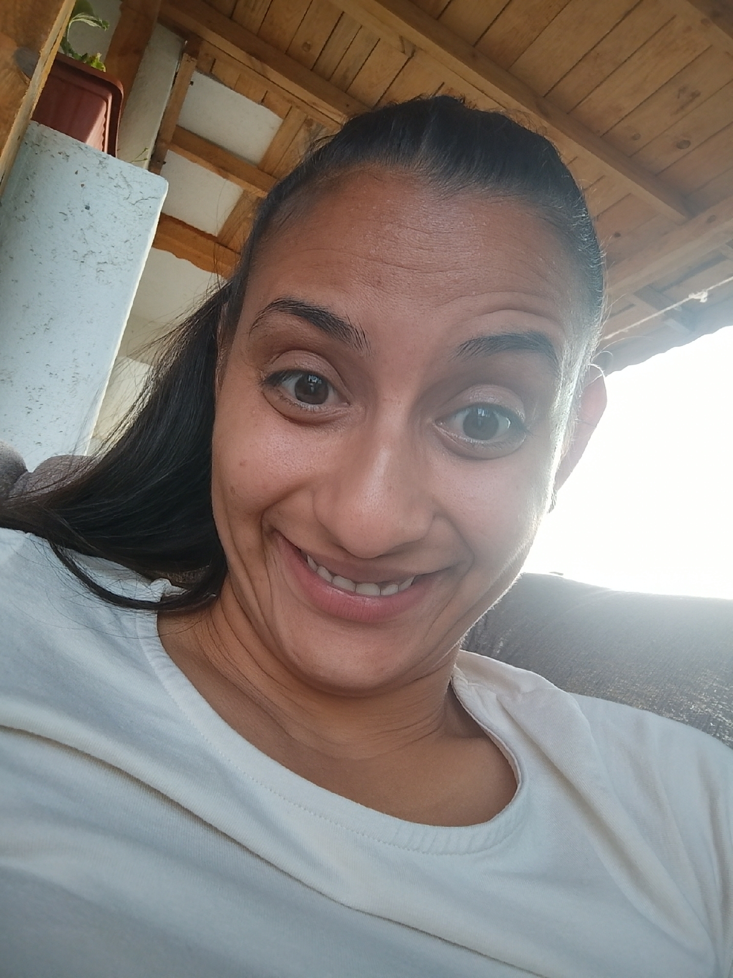 A selfie taken outdoors shows a woman with dark hair pulled back from her face, smiling broadly. She is wearing a light-colored t-shirt. The background includes wooden beams overhead and a textured white pillar to the left, suggesting an outdoor shelter or porch. A brown planter with a green plant is visible on top of the pillar. The lighting is bright, with the sun appearing to be to the right, creating a glare. The location is identified as Zimnitsa, Bulgaria.