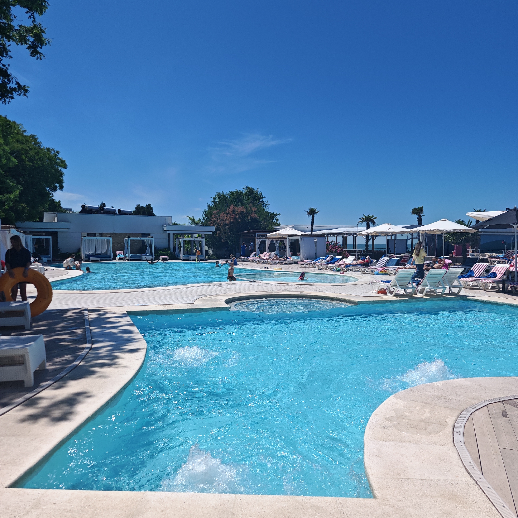 This media file depicts a lively outdoor swimming pool area on a clear, sunny day, suggesting midday or early afternoon. The scene is set in what appears to be a resort or hotel complex, with a large, sparkling blue swimming pool as the focal point. Multiple people are enjoying the water, with some swimming and others relaxing in the pool.
The pool area is well-equipped for leisure, featuring numerous lounge chairs with umbrellas, some of which are occupied by sunbathers. White cabanas are also visible, offering shaded retreats. The architecture of the buildings in the background is modern and clean, with white facades and dark window frames, further reinforcing the resort-like atmosphere. Palm trees and other lush greenery dot the landscape, adding to the tropical or vacation vibe. The bright blue sky overhead indicates pleasant weather.
Notable details include a woman in the foreground standing near the edge of the pool, holding a large orange life preserver. Children are also present, enjoying the water, and a person is seen walking along the pool deck. The water in the pool is active, with splashes and ripples indicating movement and activity. The overall mood of the scene is one of relaxation, fun, and enjoyment, typical of a vacation setting. There are no discernible text elements within the image.