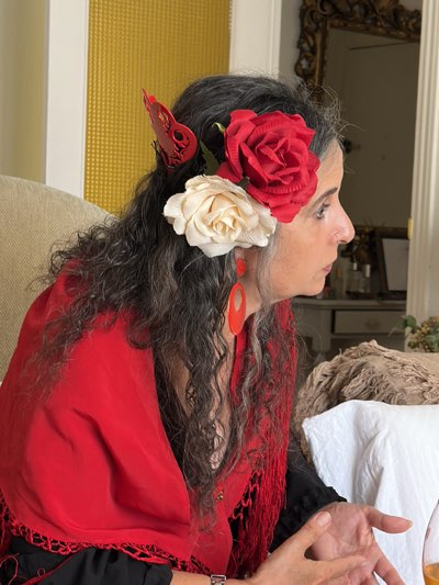 A woman with long, curly, graying dark hair is depicted in a profile view, looking intently to her right, possibly engaged in a conversation. Her hair is adorned with two large artificial flowers, one vibrant red and one cream-colored, placed above her left ear. A decorative red comb (peineta) with an intricate, possibly butterfly-like, design is visible behind the flowers. She wears large, oval-shaped red dangle earrings that match her accessories and a bright red shawl with a fringed border, draped over a black garment. Her hands are visible in the lower right, appearing to gesture.
The scene is set indoors, with a textured golden wall panel visible to the left. To the right, an ornate gold-framed mirror reflects a blurred background, suggesting a well-appointed room. The woman's attire and accessories, particularly the peineta and shawl, evoke a strong Spanish cultural aesthetic, consistent with the provided location context of Jerez de la Frontera, Spain. There is no visible text in the image.