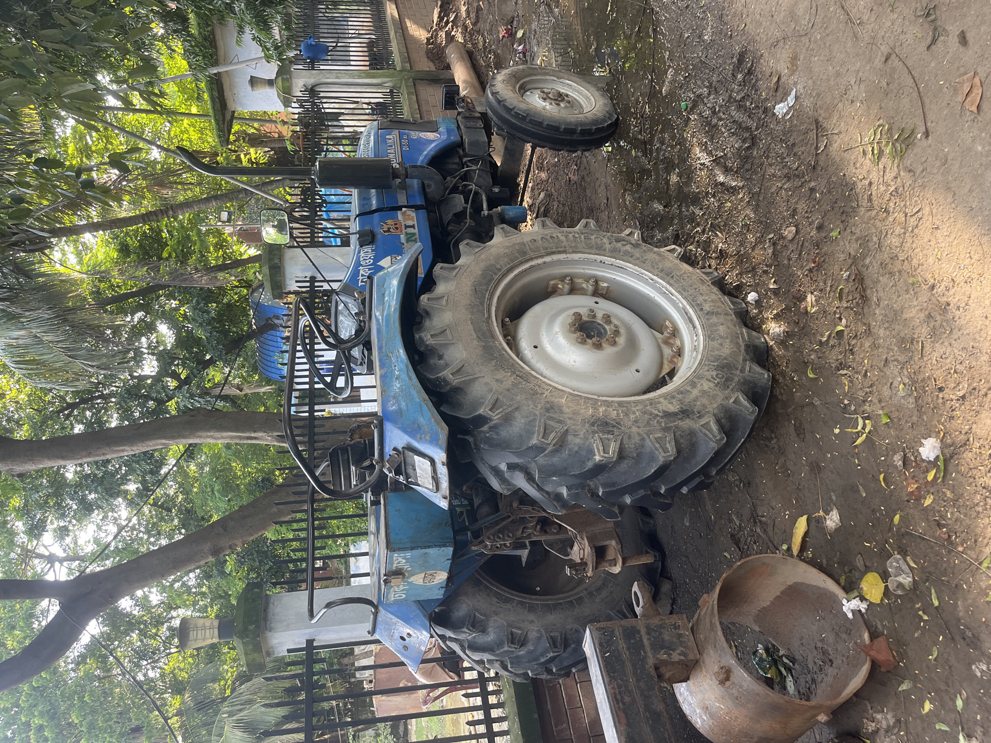 A blue Sonalika DI-50 tractor is parked on a dirt and mud road with puddles of water. The tractor has large rear tires and smaller front tires, a steering wheel, and a driver's seat. The side of the tractor has decals including "SONALIKA" and "DI-50 RX". There is text in Bengali on the side of the tractor which translates to "Dhaka Tractor". The tractor is parked next to a black metal fence. Behind the fence, there are trees and some buildings. In the foreground, there is a rusted metal cylinder and some scattered debris. The scene appears to be outdoors in a semi-urban or rural area.
