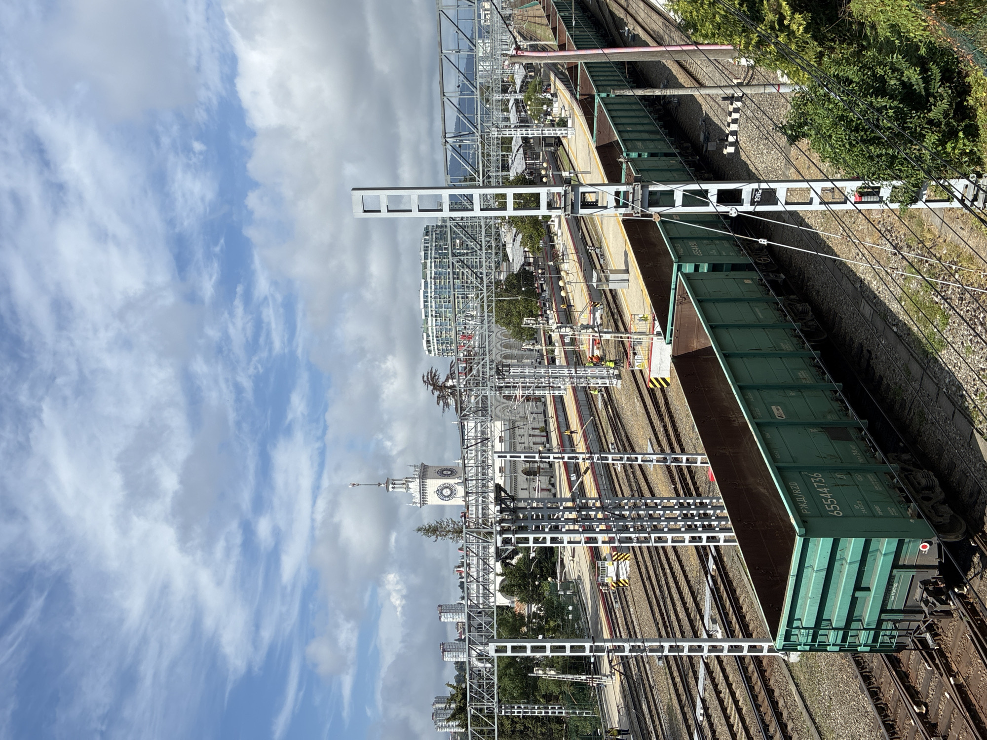 This is a high-angle, full shot of a busy train station in Sochi, Russia, on a partly cloudy day. In the foreground, a long train of green, open-top freight cars is parked on the tracks. The train cars have Cyrillic lettering and numbers visible on their sides. Beyond the freight train, multiple train tracks run in parallel, bordered by overhead power lines and support structures. Several train platforms are visible, with a few individuals in work vests appearing to be on duty. In the middle ground, a grand, white train station building with a prominent clock tower stands out. The architecture of the station is classical with arched entrances. Further in the background, modern, circular buildings and taller apartment complexes rise against the sky, hinting at the urban environment of Sochi. Lush green trees and vegetation are scattered throughout the scene, adding a touch of nature to the industrial and urban landscape. The sky is a mix of blue patches and white, scattered clouds.