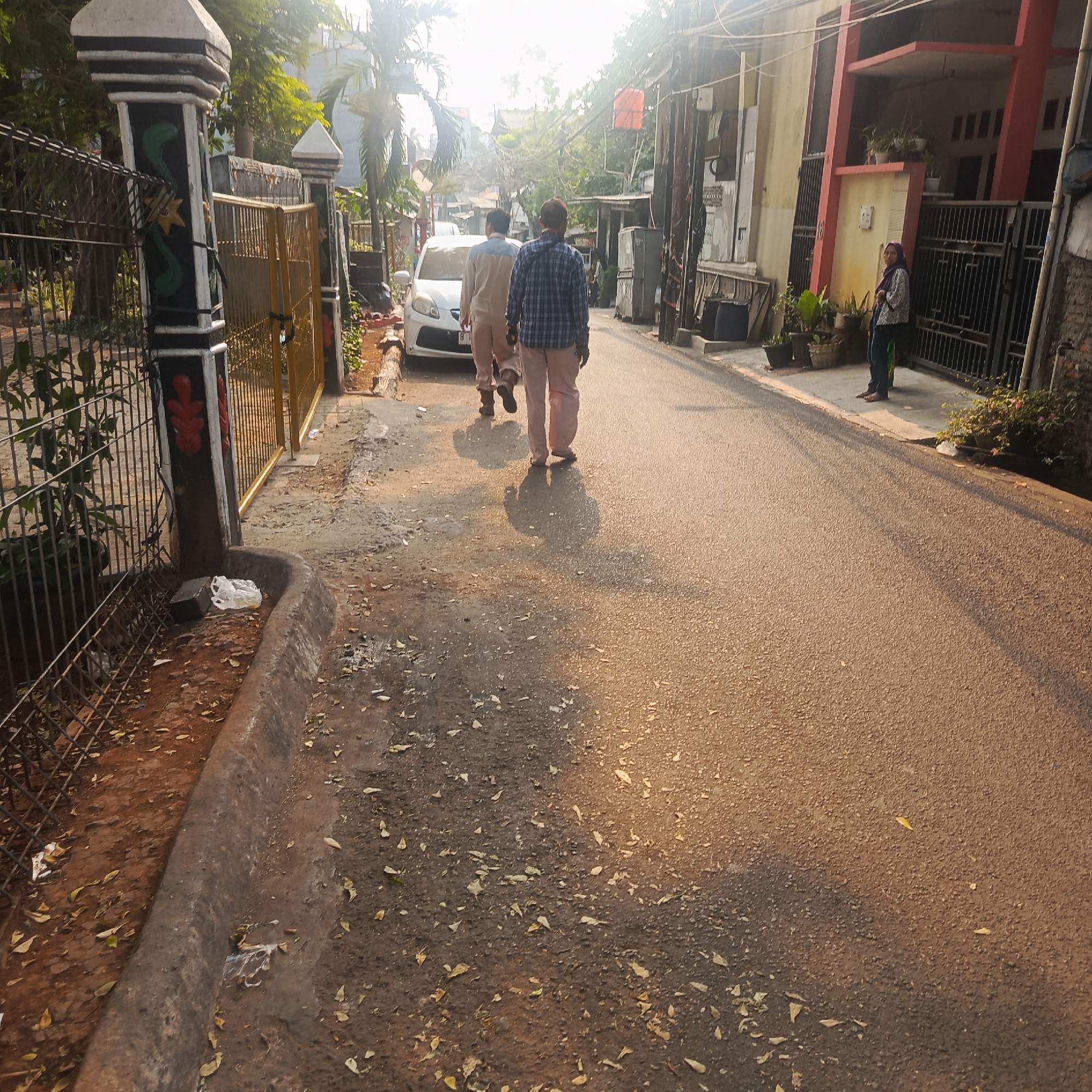 The image captures a sunlit residential street in South Jakarta, Indonesia. Two men walk away from the camera down the asphalt road, casting long shadows. The man in the foreground wears a blue and white plaid shirt and light-colored pants, while the man behind him wears a tan shirt and similar light-colored pants. To the left of the street is a metal fence with decorative pillars, one of which displays painted red and green motifs. Beyond the fence, a yellow gate is visible. A white car is parked on the left side of the street. The right side of the street is lined with houses, one with a red-accented second story and black metal gates. A woman wearing a headscarf and patterned shirt stands near the gate of one of the houses. Scattered leaves litter the road and the area beside it. The overall impression is of a typical, somewhat cluttered urban street scene bathed in the warm light of a sunny day.