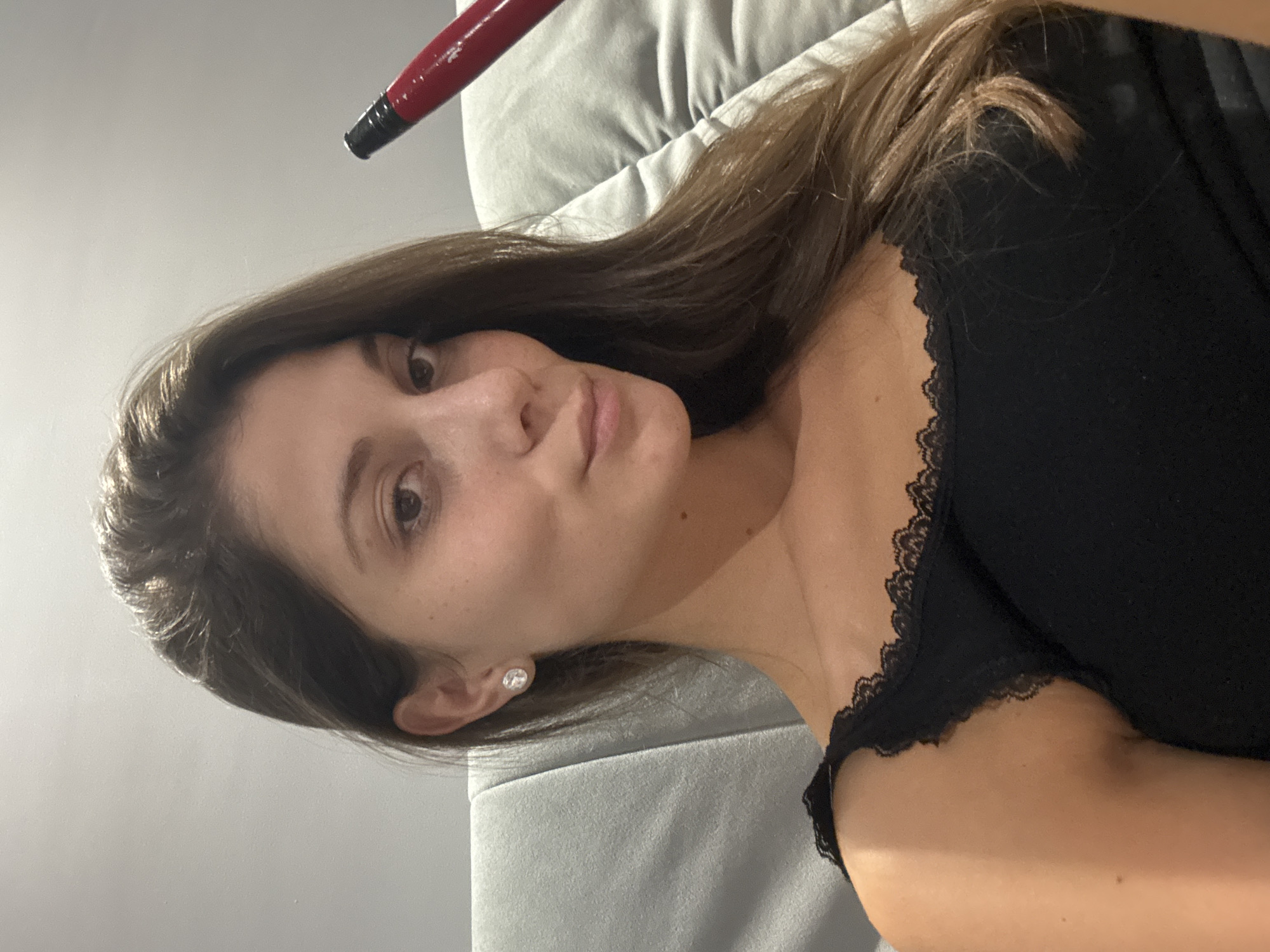 The image shows a medium close-up of a young woman with long, dark brown hair, wearing a black tank top with lace trim. She has fair skin, brown eyes, and is wearing a stud earring in her left ear. She is holding a red object, which appears to be a hookah hose, to the right side of her face. She is seated on a grey couch. The background is a plain, light-colored wall. There is no visible indication of the location being in the city of Burgas, Bulgaria.