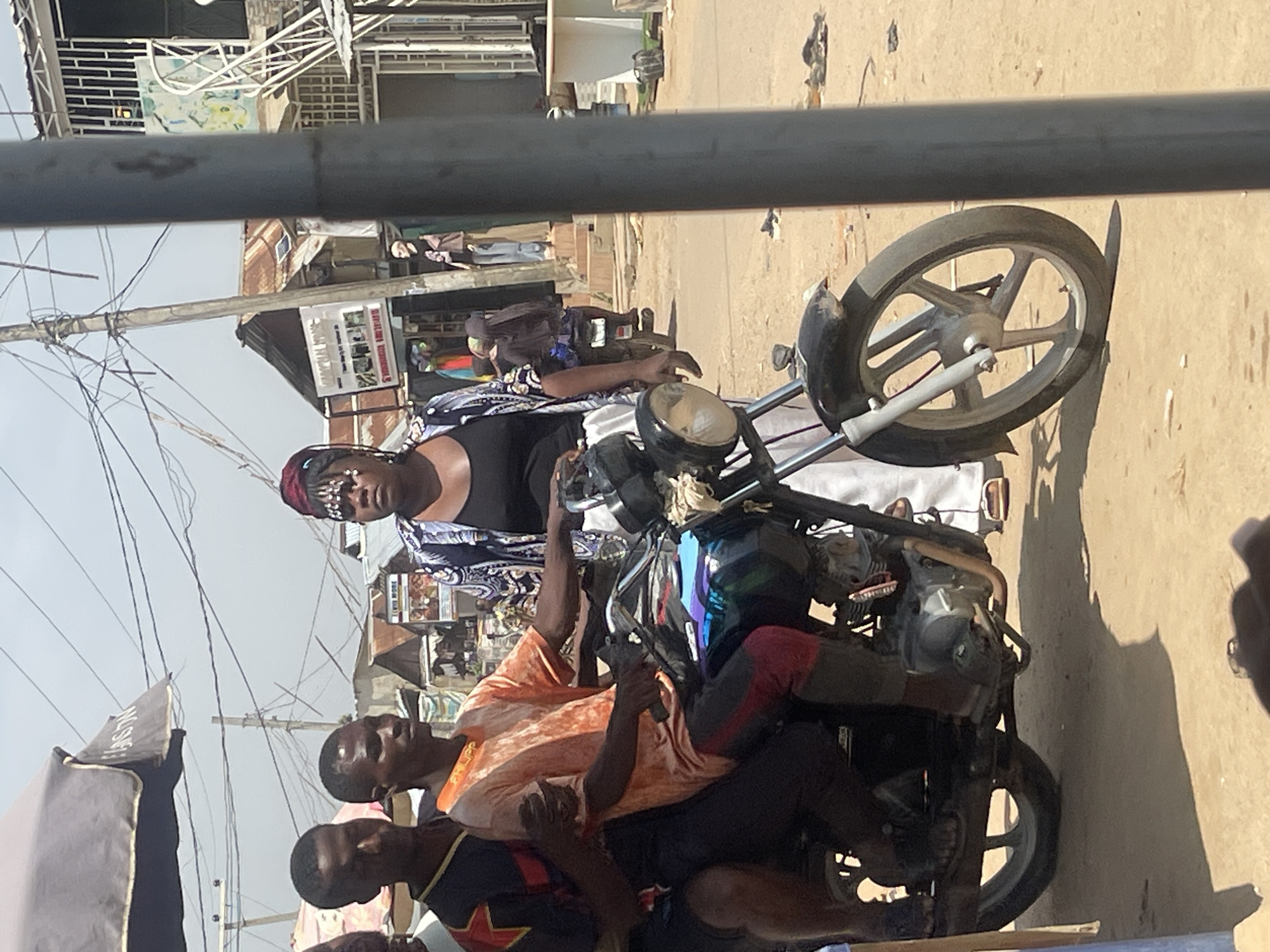 The image depicts a street scene in Abuja, Nigeria, on a bright, sunny day. In the foreground, a motorcycle is the central focus, carrying three people. The driver, an adult male wearing an orange t-shirt with the word "PHILIPP" printed on it in yellow, is seated at the front. To his left and behind him sits another adult male wearing a black and red patterned shirt. A third person, an adult female with elaborate beaded hair ornaments and distinctive makeup, is seated behind the driver, facing forward. She is wearing a black tank top and a patterned open-front jacket over it, paired with a white skirt. The motorcycle itself is dark colored, with a prominent front wheel and engine visible.
The background reveals a bustling street environment. Several market stalls or shops are visible, characterized by corrugated metal roofs and various goods displayed. A prominent billboard or sign with text and images is mounted on a pole. Power lines crisscross the sky, indicative of urban infrastructure. In the distance, more buildings and a mannequin standing on steps can be observed, suggesting a commercial area. The ground is unpaved, covered in sand or dirt, with scattered debris. The overall impression is one of everyday life and activity in a Nigerian city.